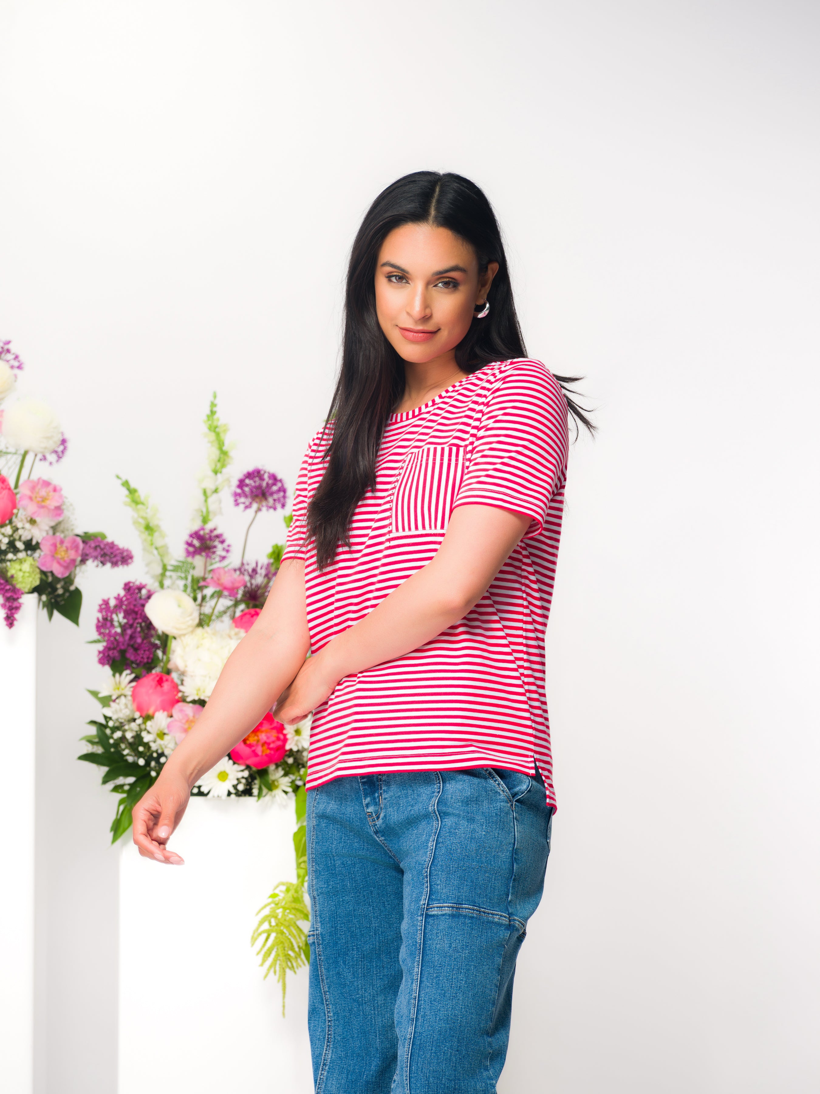 Model wearing a red and white striped short sleeve cotton tee with chest pocket, styled with blue jeans, studio background with floral arrangement.