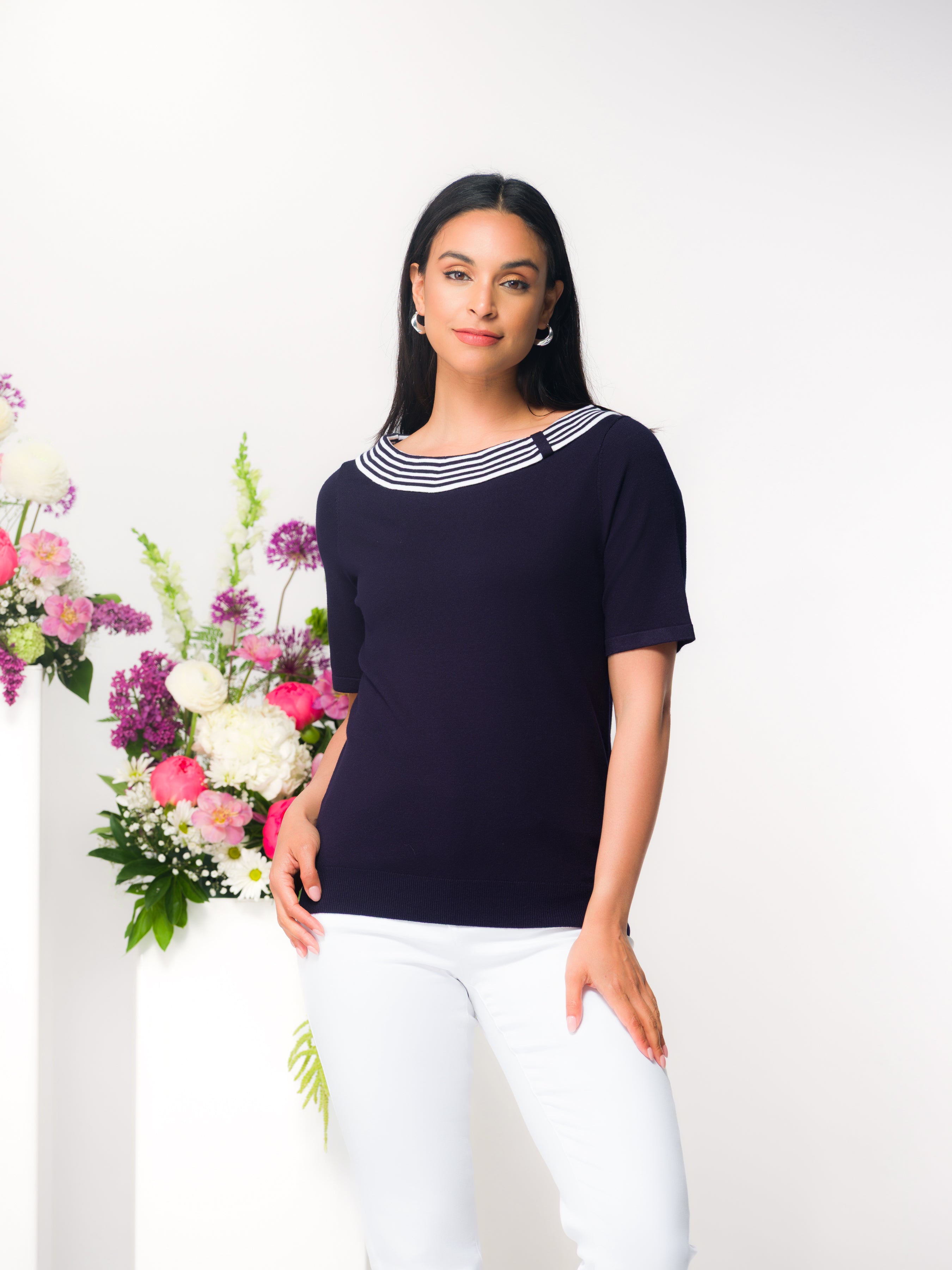 Model standing in a studio wearing a navy short-sleeve knit sweater with a striped bateau neckline and white slim-fit pants, posed beside a white pedestal with a pink and white floral arrangement.