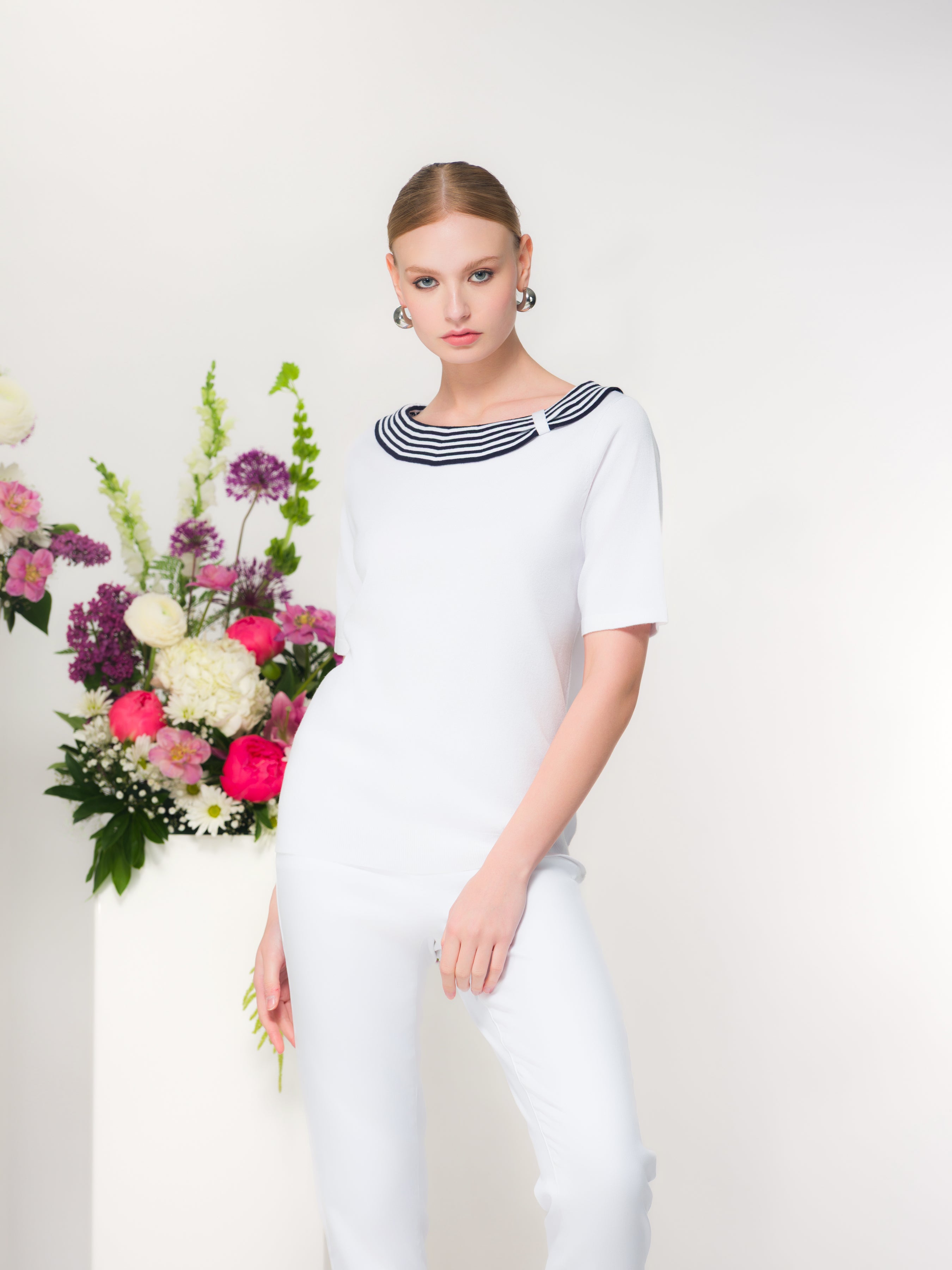 Model standing in a studio wearing a white short-sleeve knit sweater with a striped bateau neckline and matching white slim-fit pants, posed beside a pedestal with a pink and white floral arrangement.