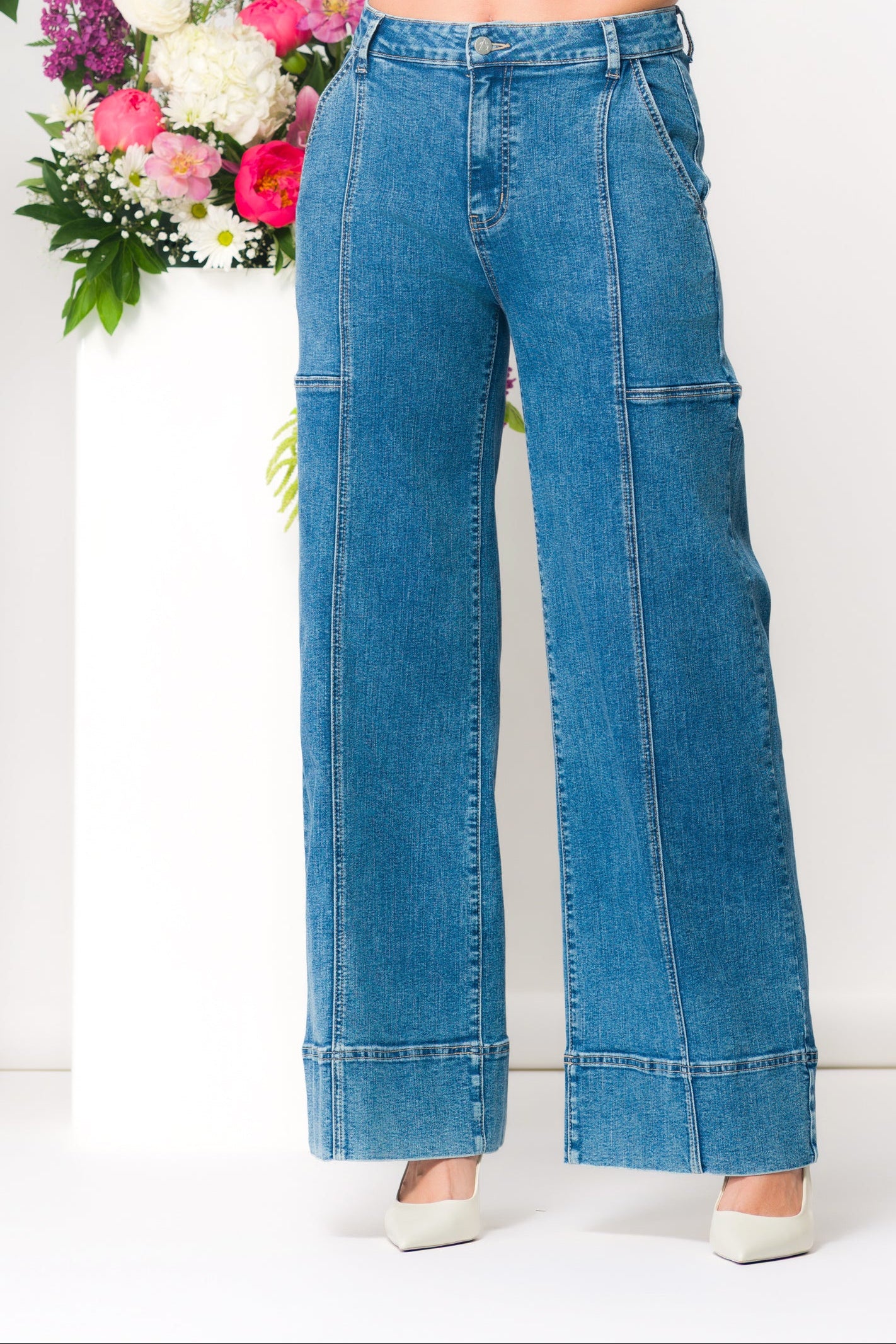 Woman modelling high-rise wide-leg denim pants with front seam detailing and cuffed hems, paired with a long-sleeve white cropped blouse and pointed-toe heels, standing beside floral arrangements against a light neutral background.