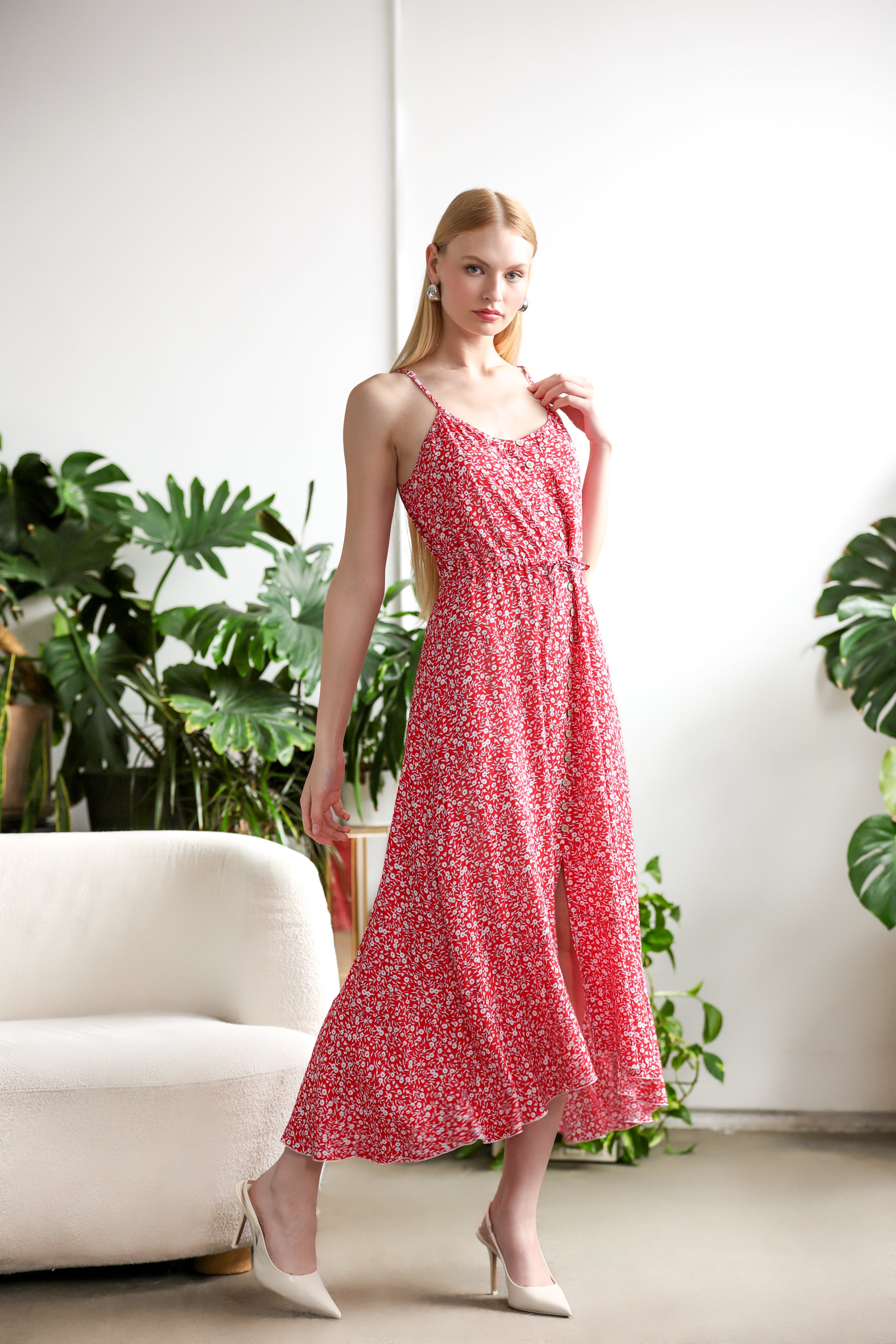 Model wearing a red floral midi dress with button-front detail, adjustable straps, and front slit, styled with cream pointed heels in a bright indoor setting with green plants.