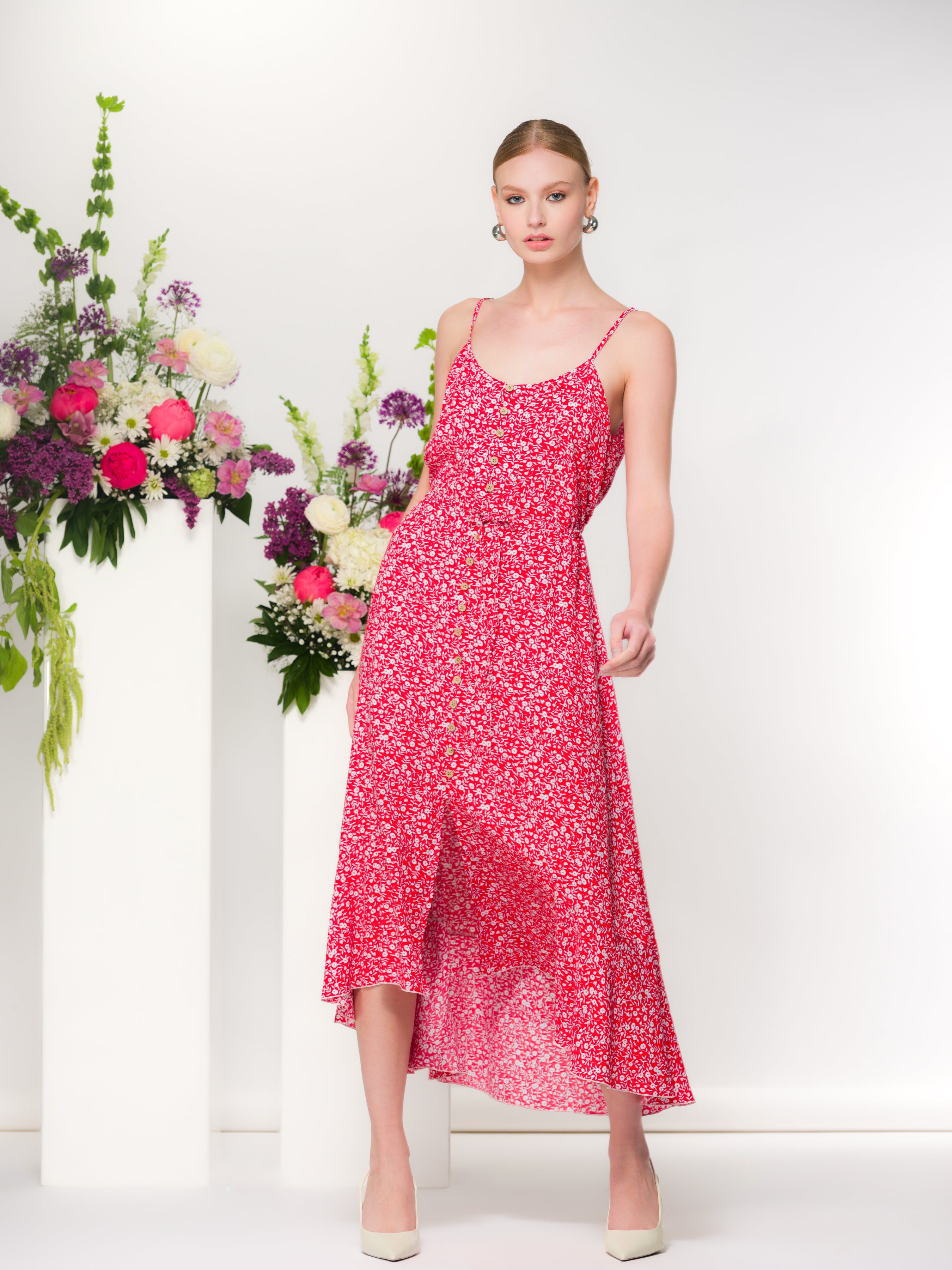 Front view of red floral button-front midi dress with adjustable straps and flowy silhouette, paired with cream heels beside a pedestal of fresh flowers.
