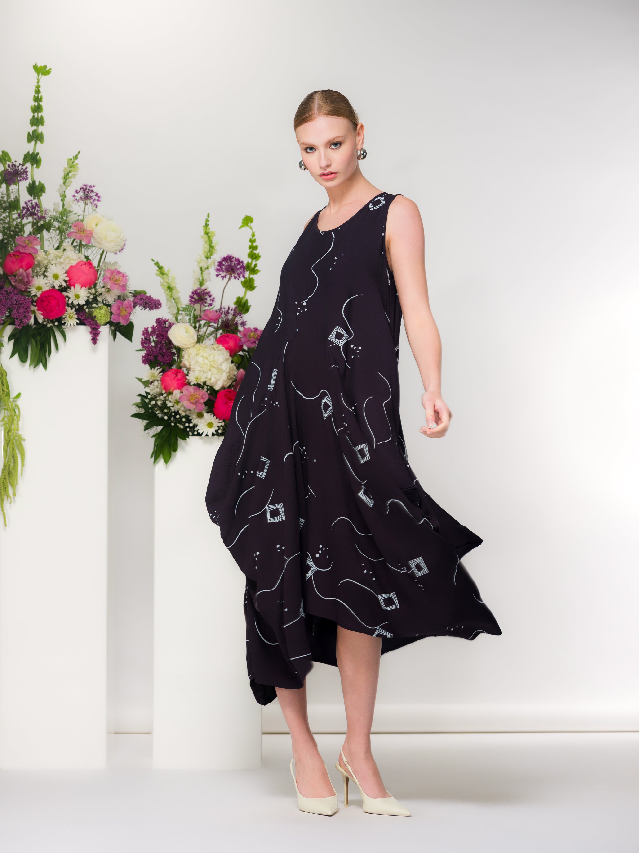 Model wearing a navy sleeveless midi dress with abstract white print and flowing asymmetrical hem, paired with cream pointed heels, photographed beside a white pedestal with colourful floral arrangement.