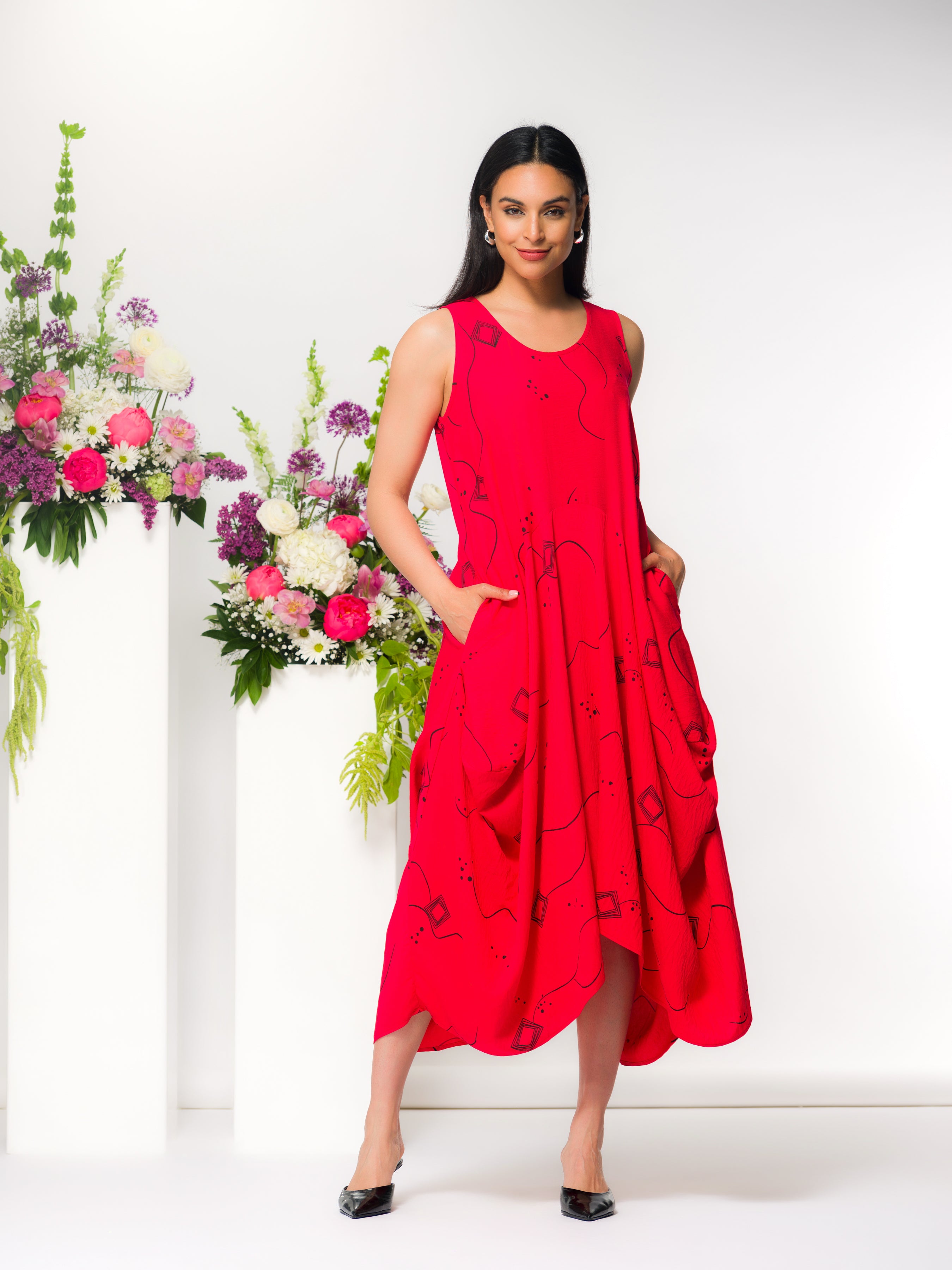 Model wearing a bright red sleeveless midi dress with black abstract print, relaxed draped fit and side pockets, styled with black pointed heels, posed next to a white pedestal with pink and white flowers.