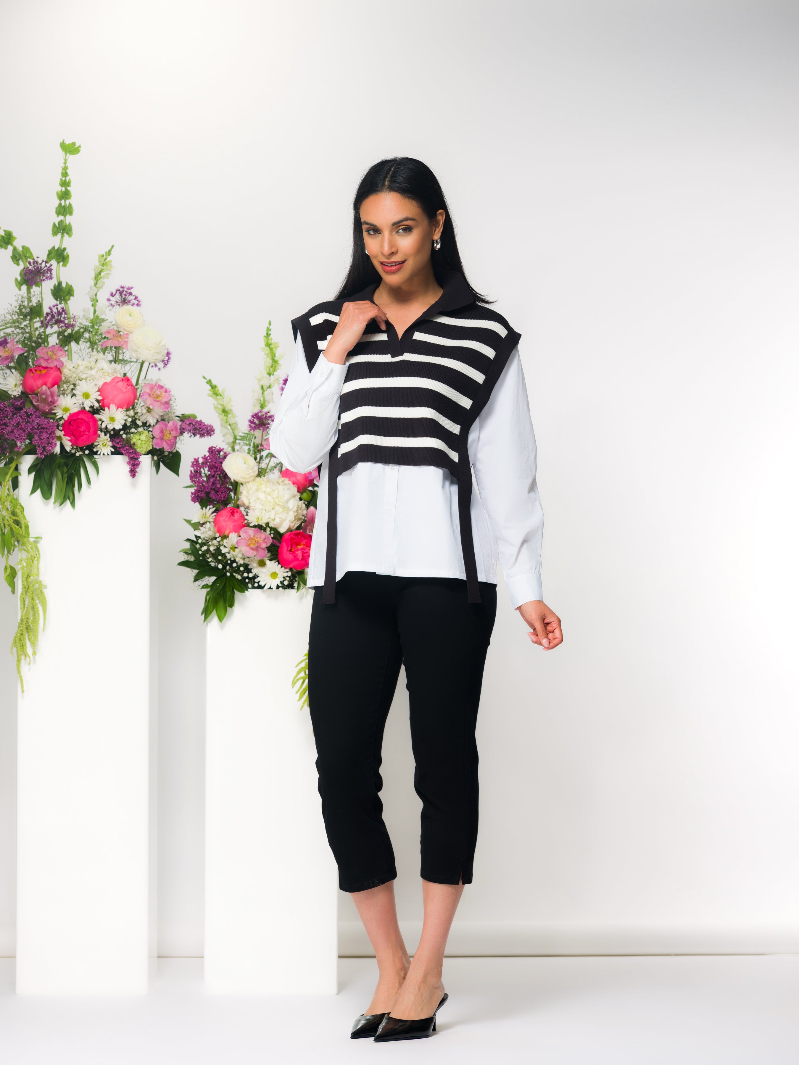 Woman modelling a layered sweater top featuring a black and white striped knit vest over a white collared shirt, paired with slim black ankle-length pants and black pointed flats, standing beside white pedestals with pink and white floral arrangements against a light neutral background.
