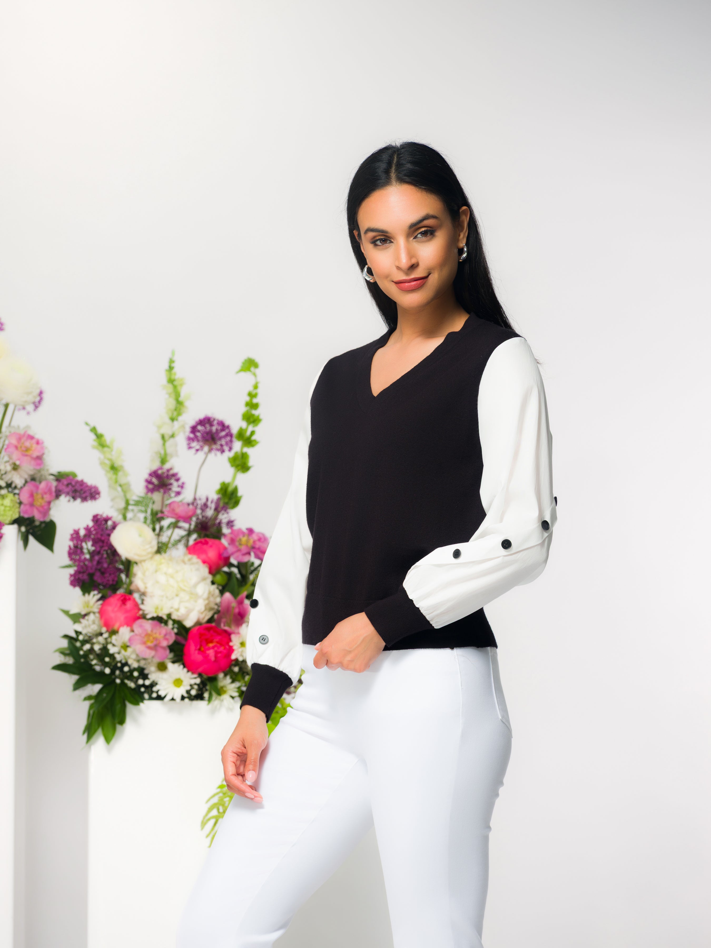 Woman modelling a two-tone cotton sweater with a black knit body and white woven button-detail sleeves, styled with fitted white pants, standing beside a floral arrangement against a clean studio background.