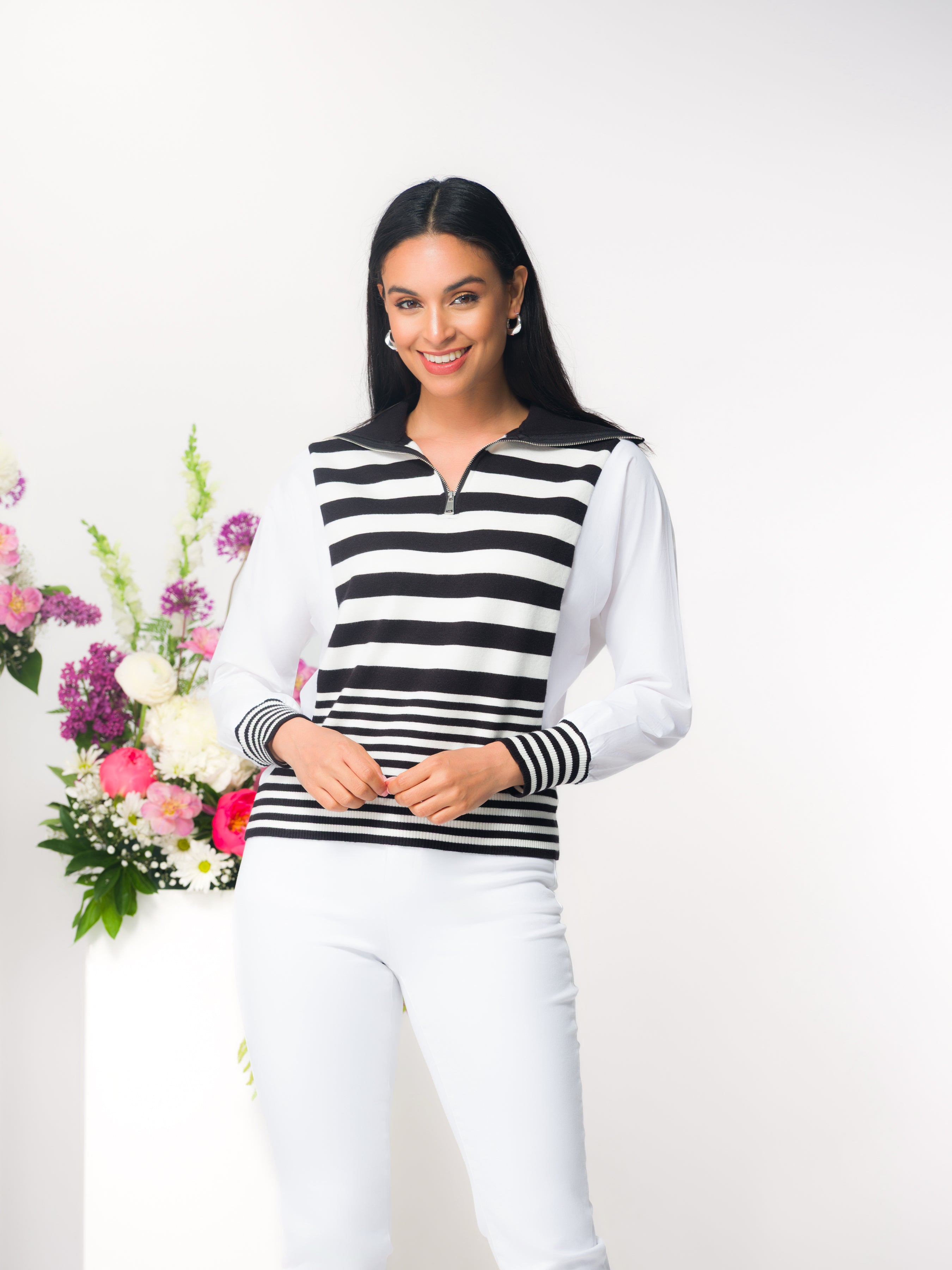 Smiling woman wearing a black and white striped half-zip knit sweater with a collared neckline and long white sleeves, paired with fitted white pants, standing beside a floral arrangement against a neutral background.