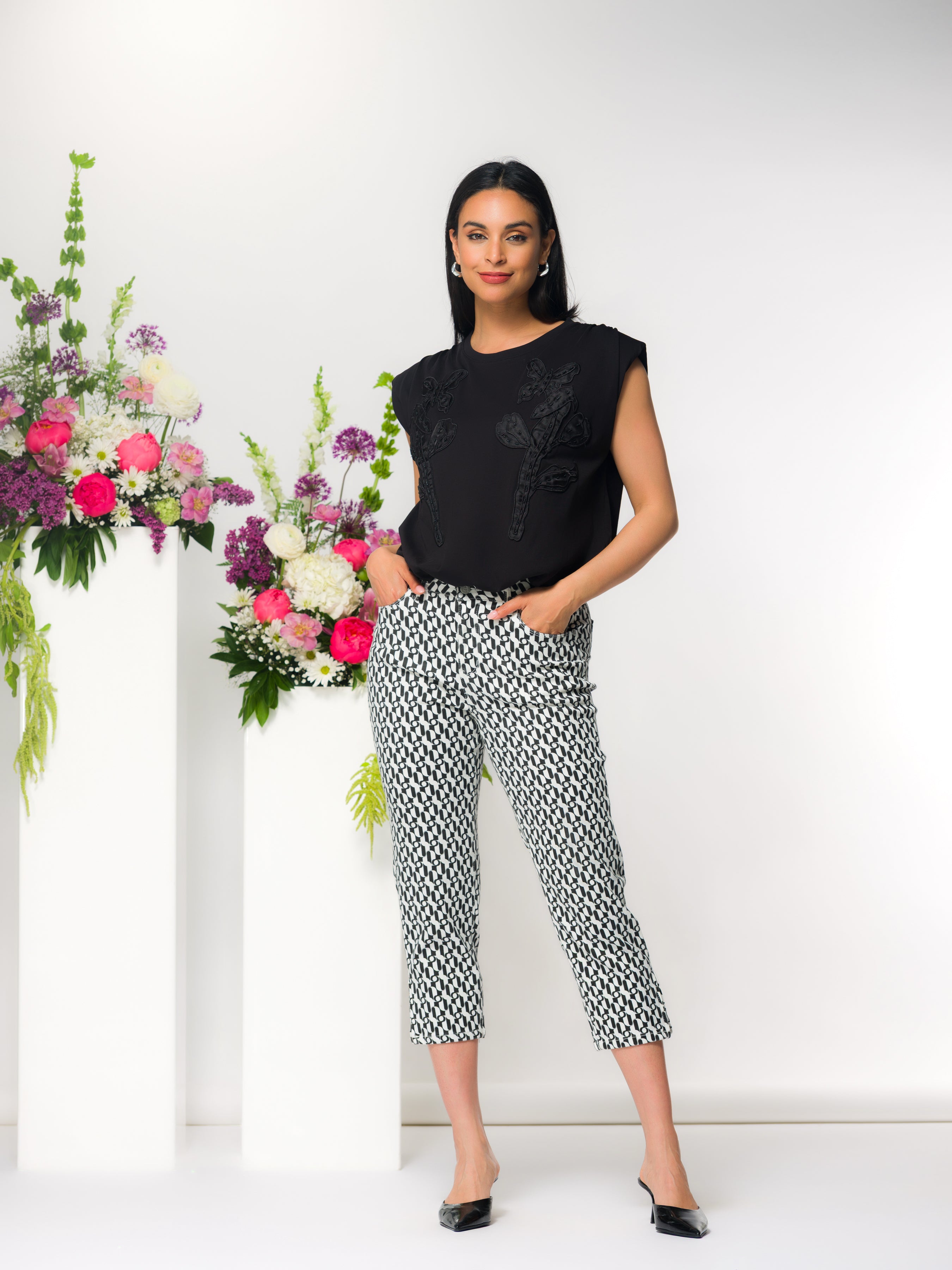 Woman wearing a black sleeveless embroidered top with a relaxed fit, paired with cropped patterned trousers and pointed flats, standing beside a floral arrangement in a bright studio setting.