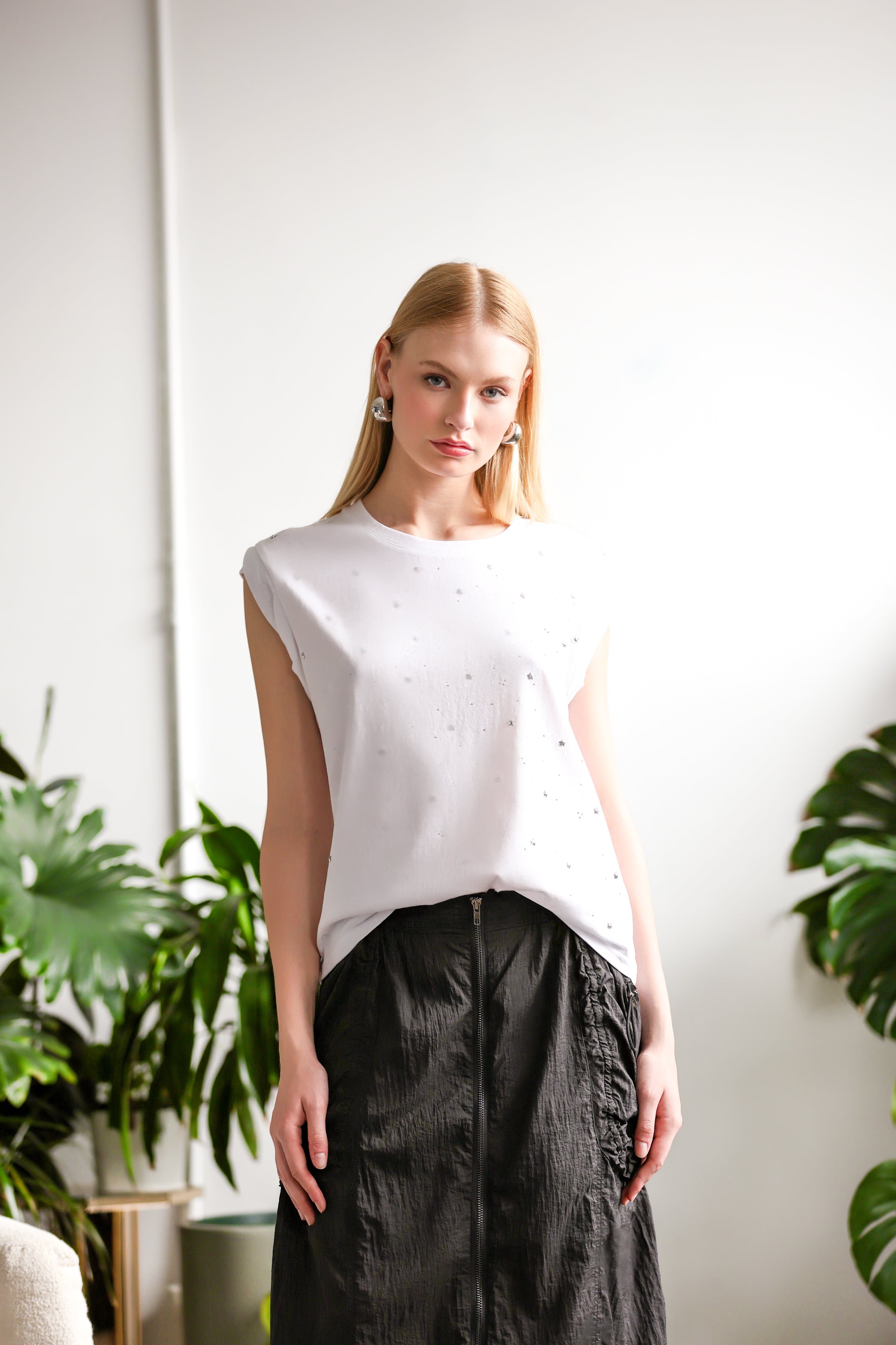 Woman wearing a white sleeveless t-shirt with subtle texture, paired with a black midi skirt and silver heeled sandals, standing in a bright indoor space with green plants.