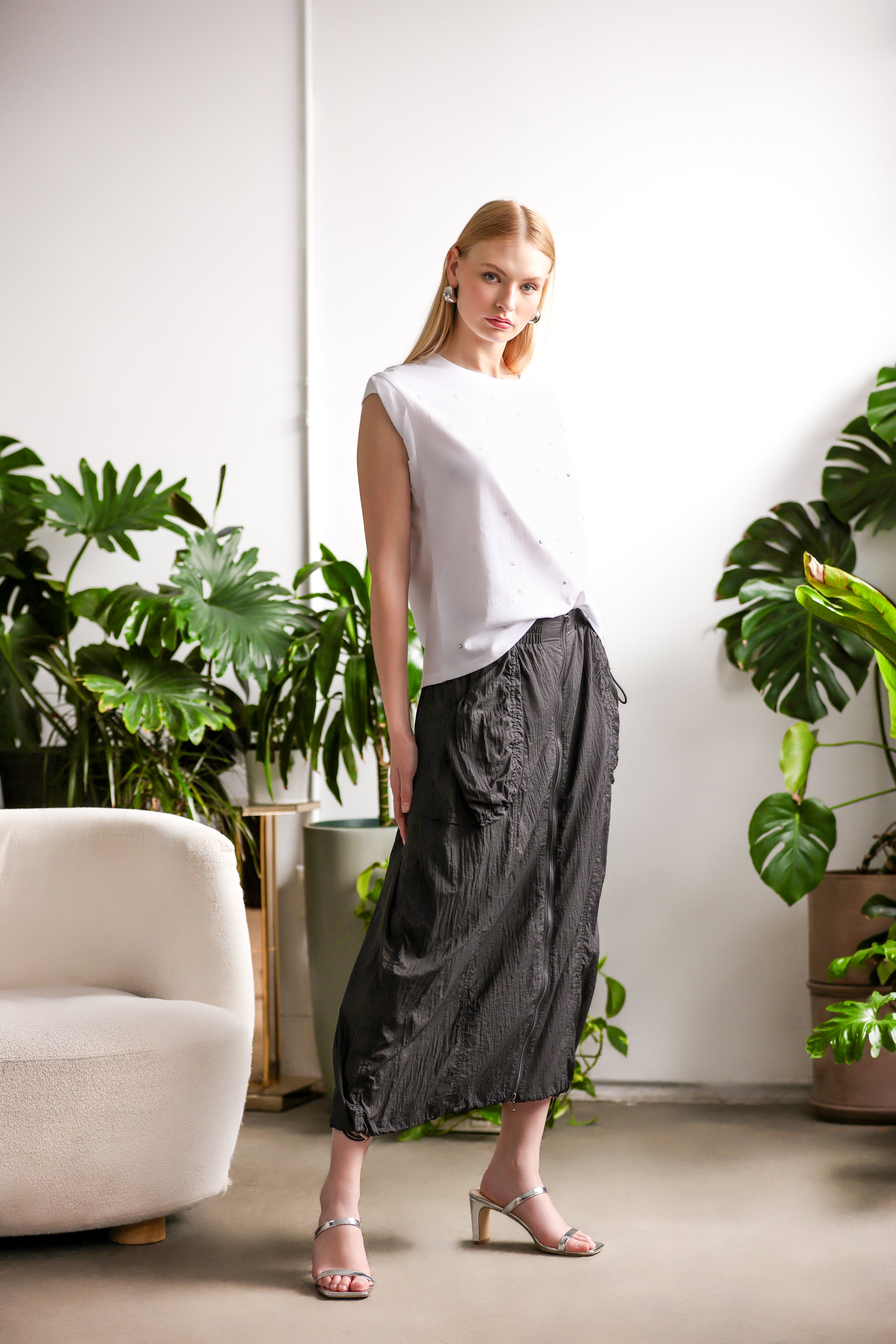 Woman wearing a white sleeveless t-shirt with subtle texture, paired with a black midi skirt and silver heeled sandals, standing in a bright indoor space with green plants.