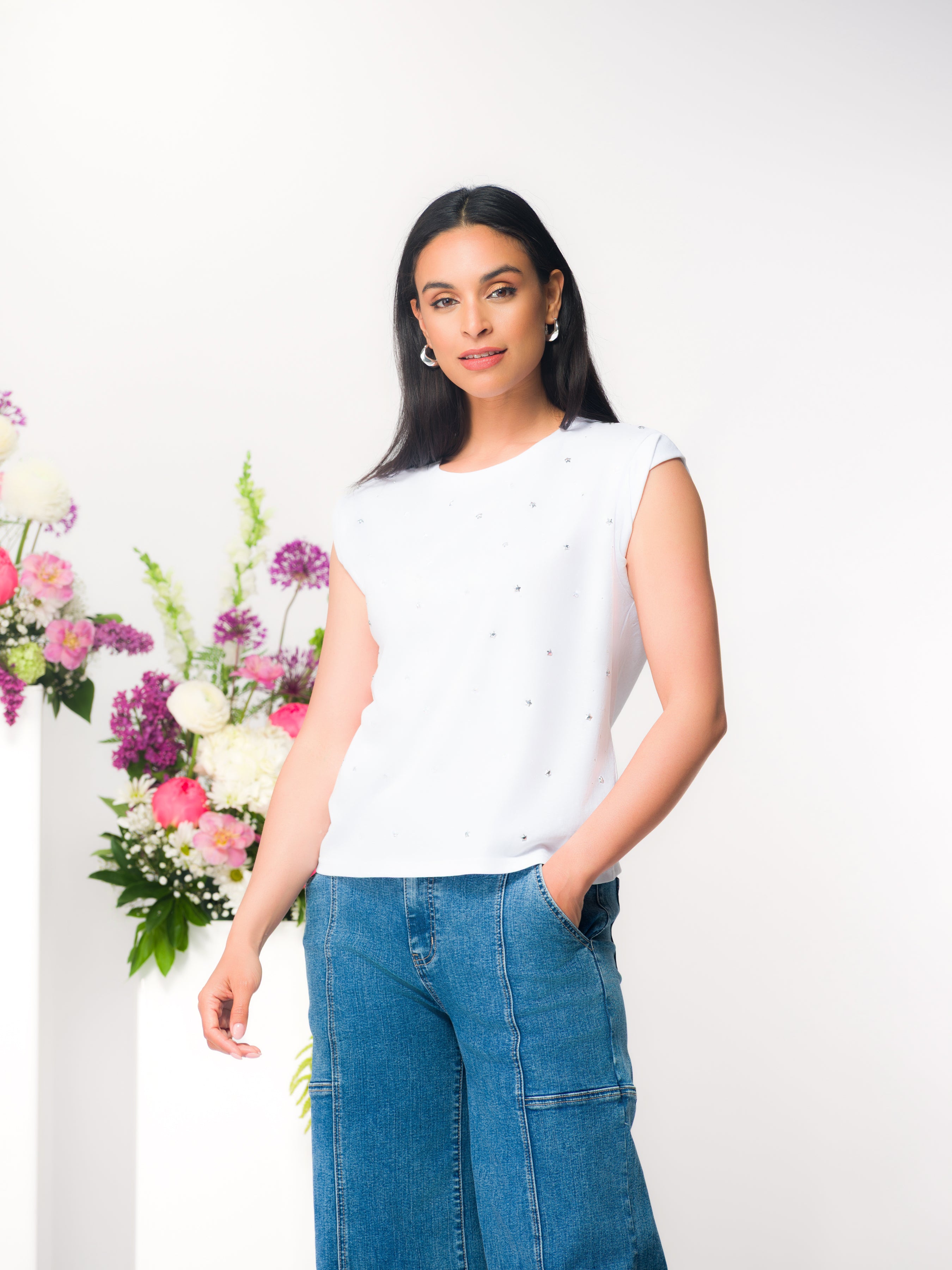 Woman wearing a white sleeveless t-shirt with delicate textured detailing, styled with blue jeans, standing against a light background with floral accents.