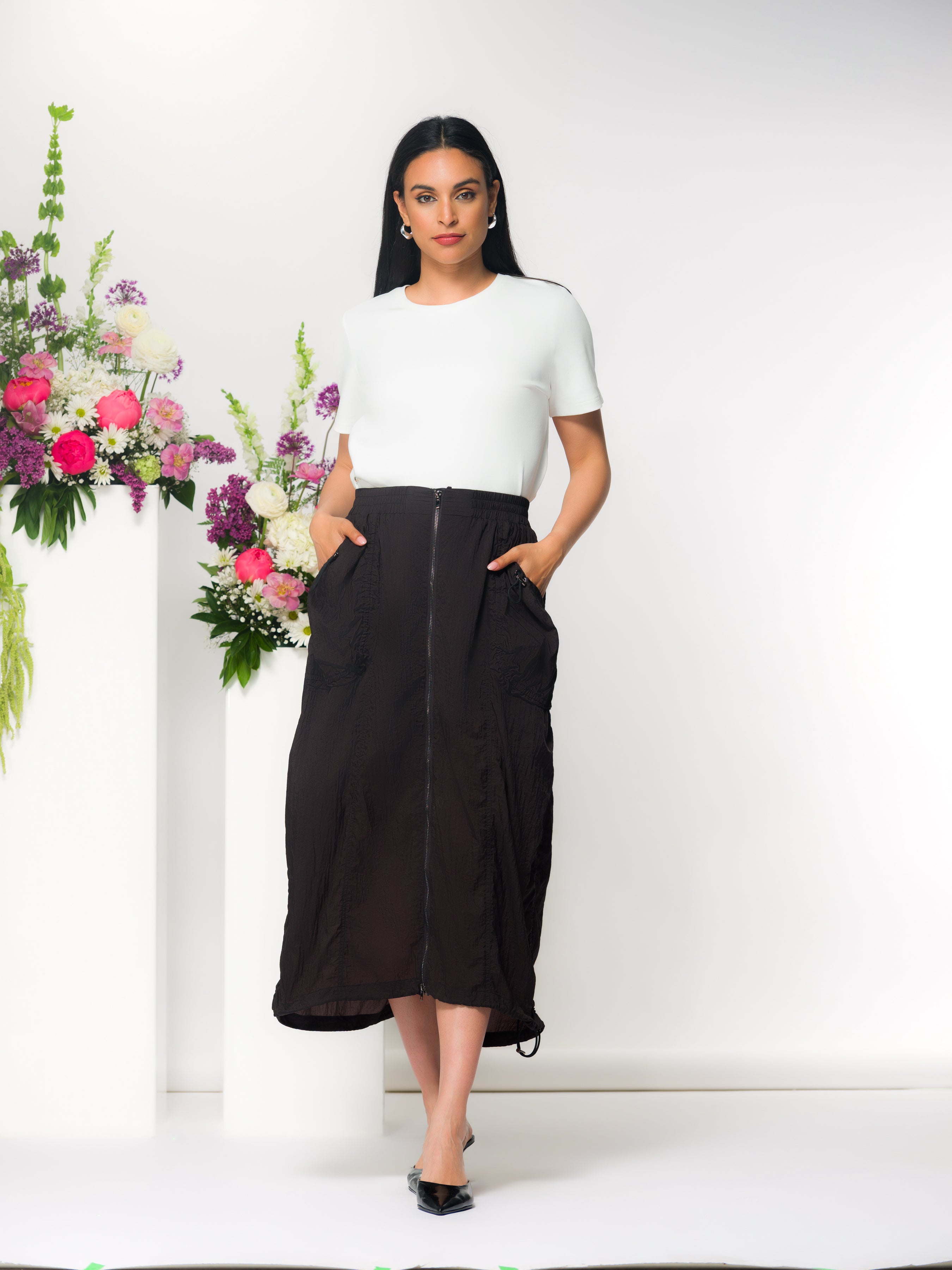 Woman wearing a white short-sleeve top paired with a black midi skirt featuring a front zip closure and side pockets, styled with pointed flats, standing against a clean studio backdrop with floral arrangements.