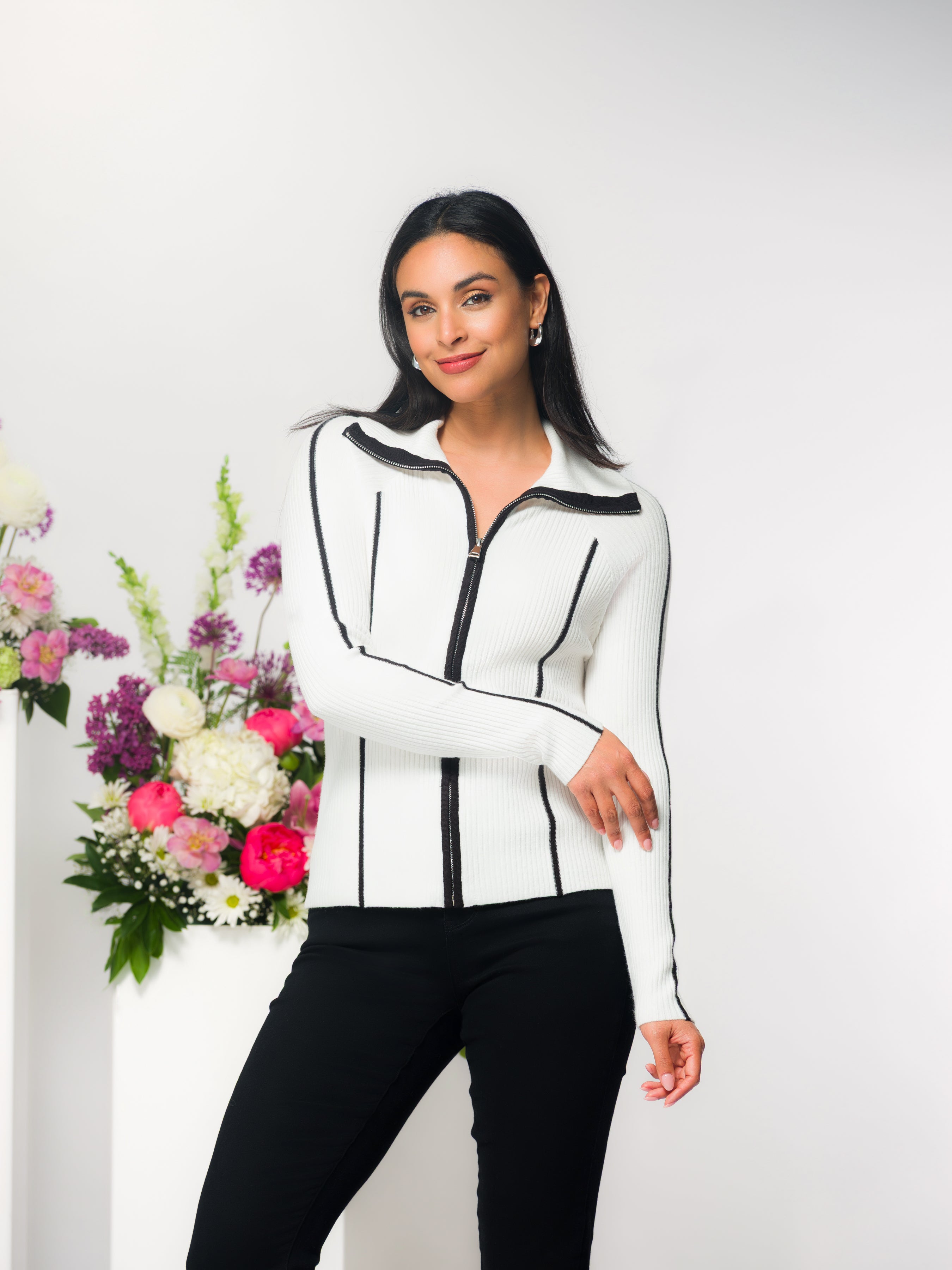 Woman wearing a cream ribbed zip-front cardigan with black vertical stripe detailing, styled with black fitted pants, standing against a neutral backdrop with a floral arrangement.
