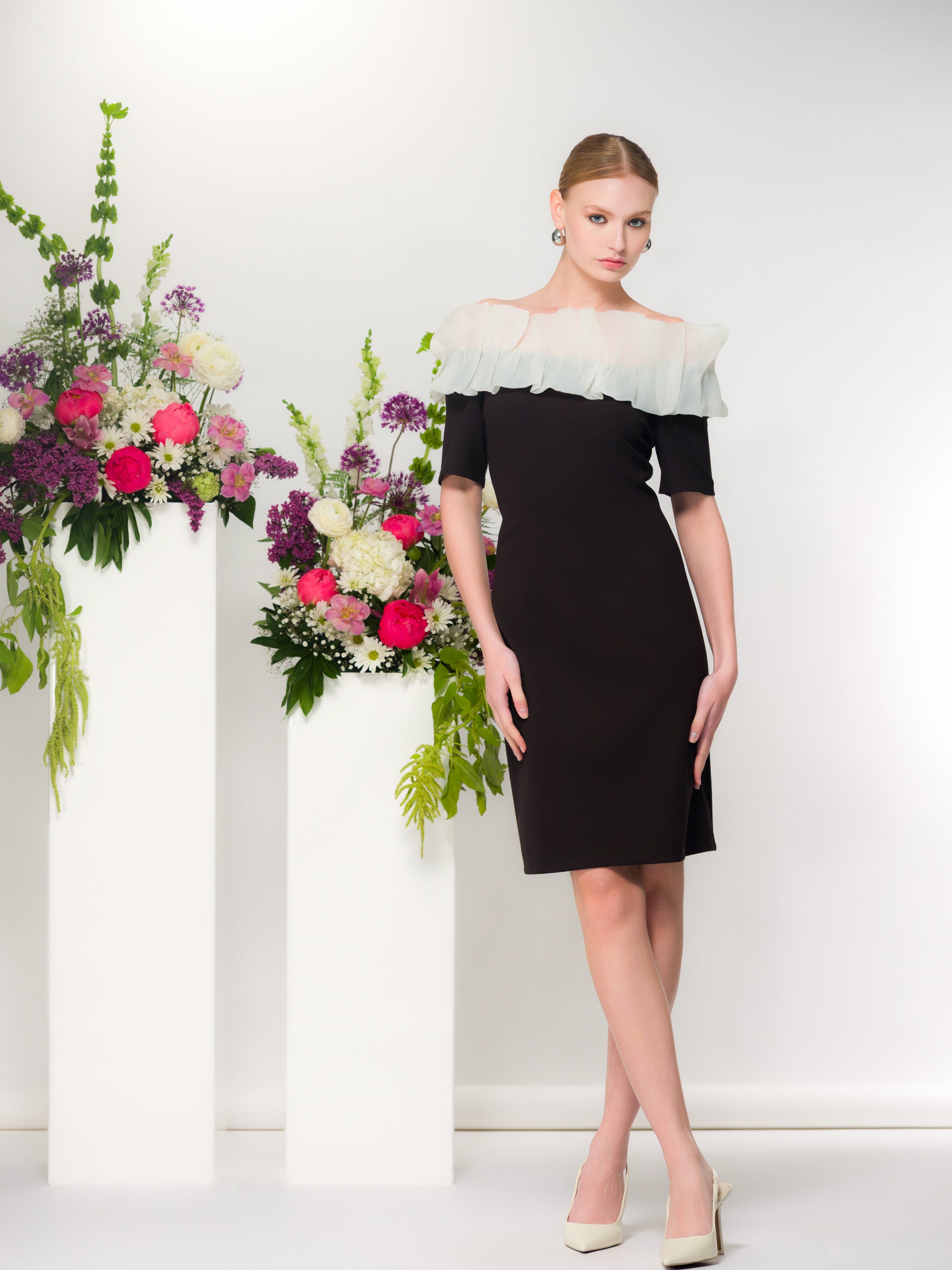 Model wearing a black knee-length sheath dress with a sheer white ruffle neckline and elbow-length sleeves, styled with nude pointed heels, standing beside tall white pedestals topped with pink and white floral arrangements.