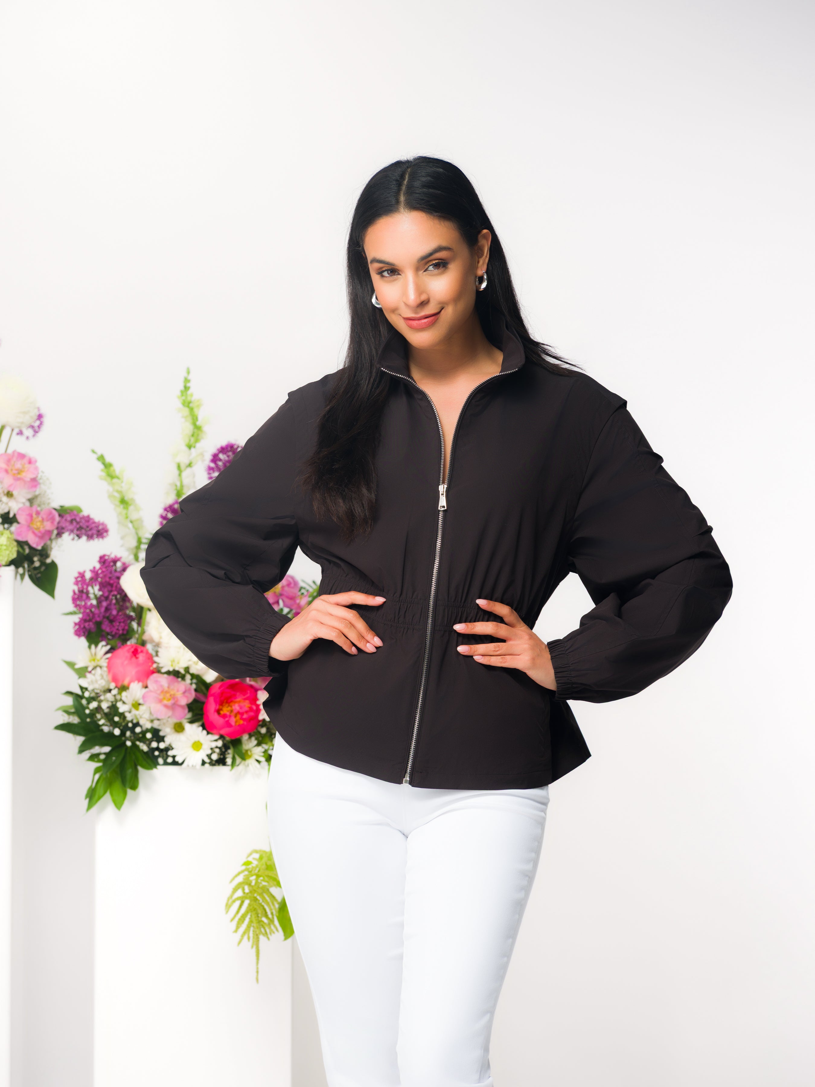 Woman modelling a black zip-front jacket with a softly gathered waist and relaxed fit, paired with light grey slim pants, standing in a studio beside a floral arrangement.
