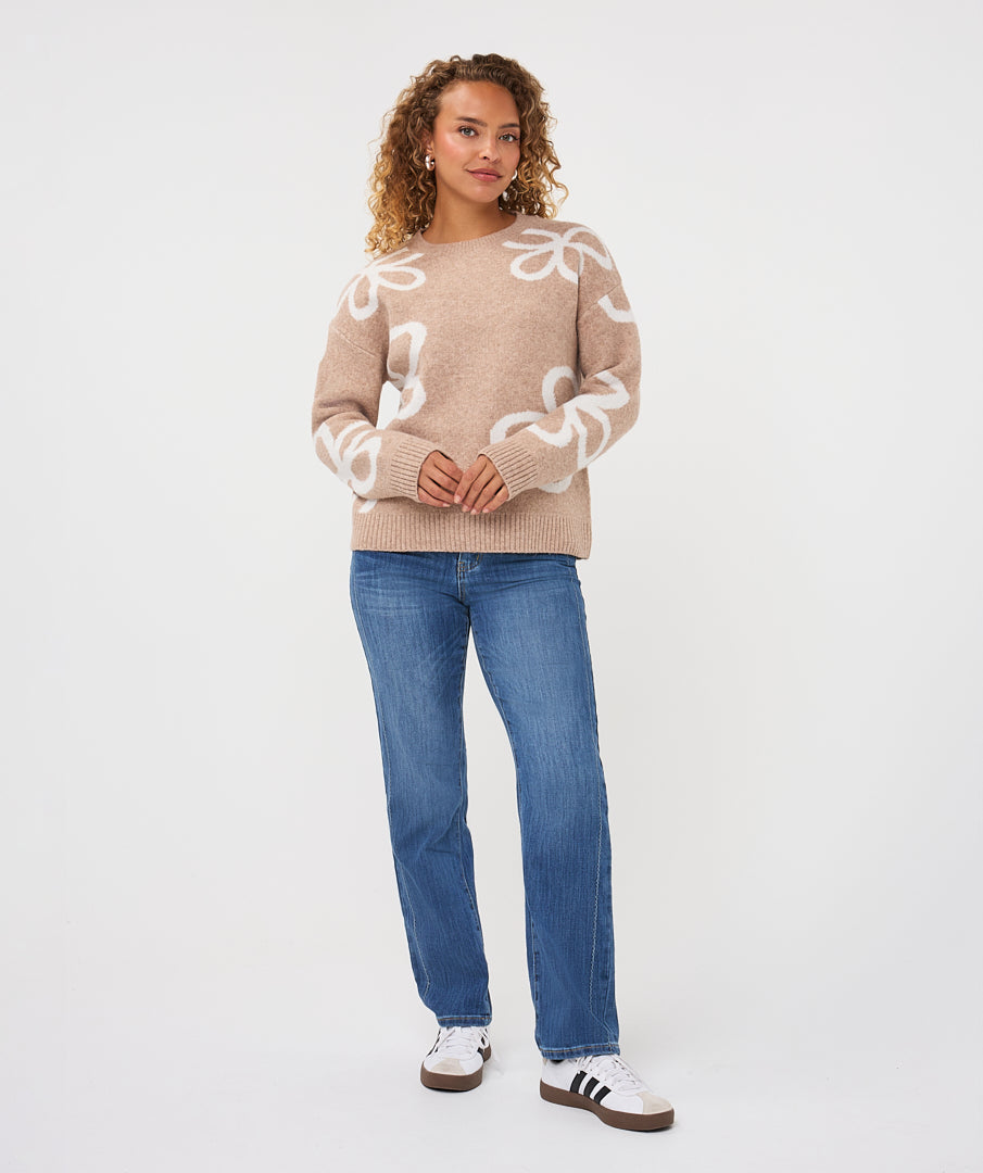 Woman wearing a sand patterned crew-neck sweater with straight-leg jeans and casual sneakers, standing against a light studio background.