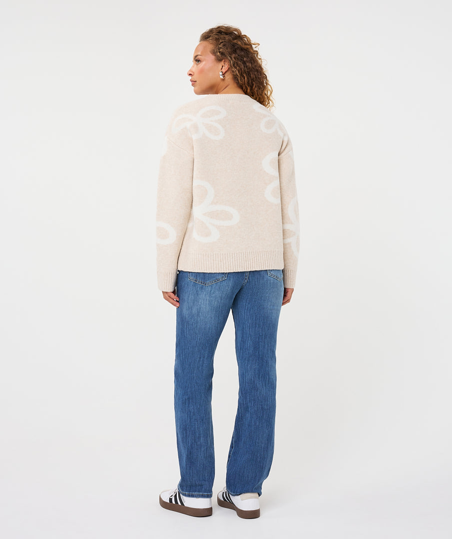 Back view of a woman wearing a beige patterned crew-neck sweater with straight-leg jeans and casual sneakers, shown in a relaxed standing pose against a light studio background.