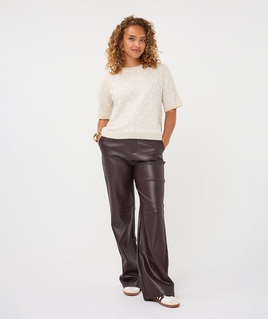 Woman wearing the cream Rosalie Lace Top with dark brown leather-look wide-leg trousers and casual sneakers, front view.