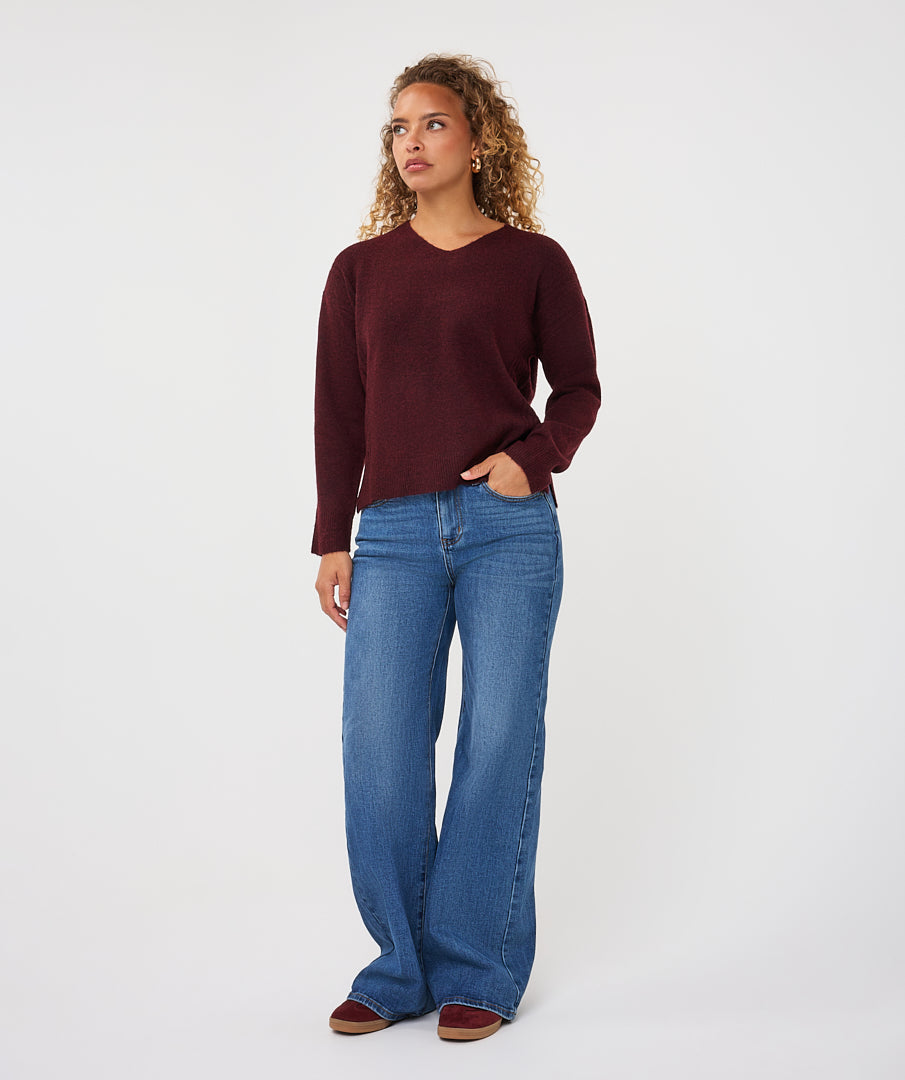 Woman wearing a burgundy V-neck sweater with long sleeves and a relaxed fit, paired with wide-leg blue jeans.