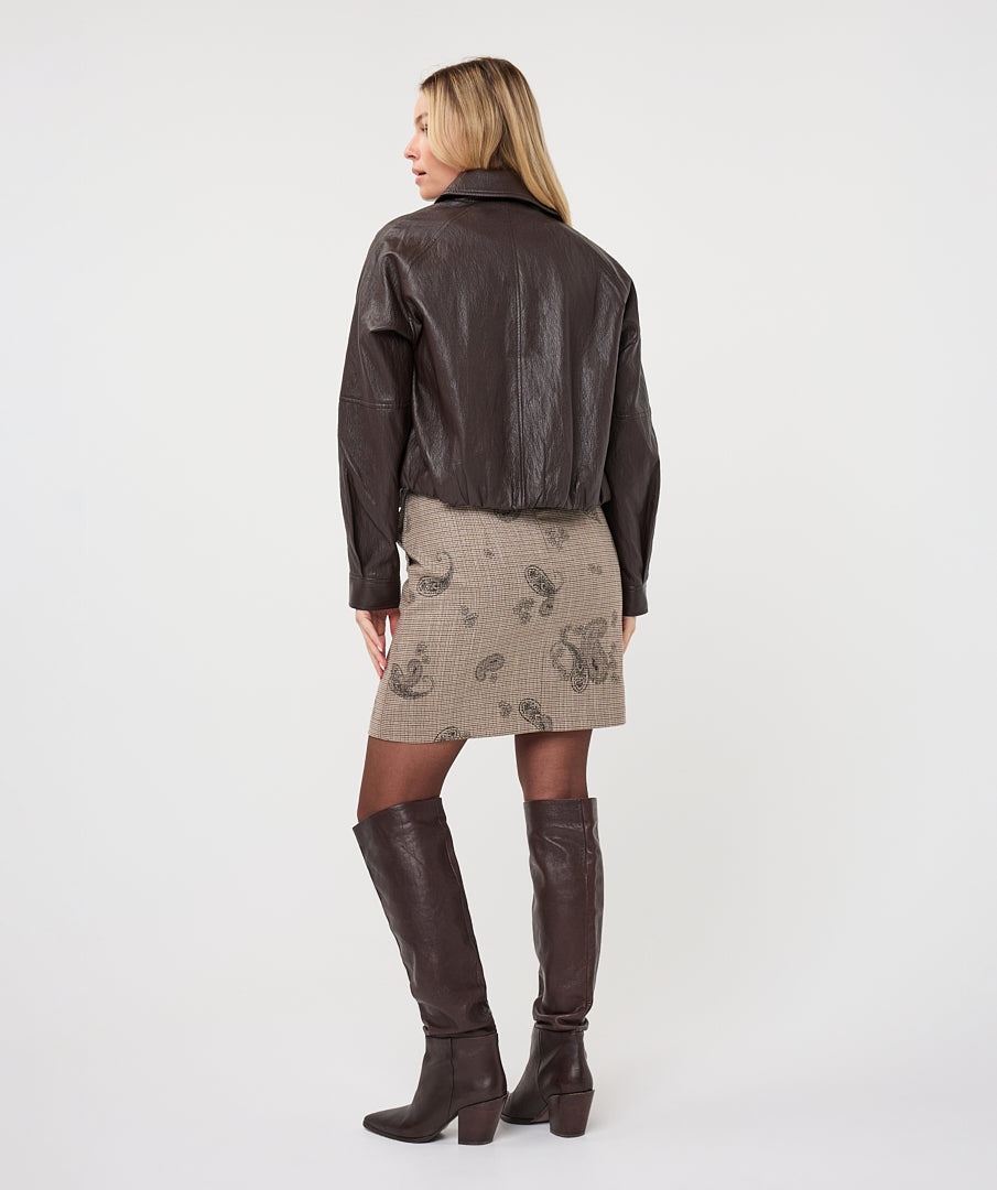 Woman wearing a dark brown cropped faux leather jacket with a collared neckline and button-front closure, styled with a brown knit top, patterned mini skirt, tights, and knee-high boots.
