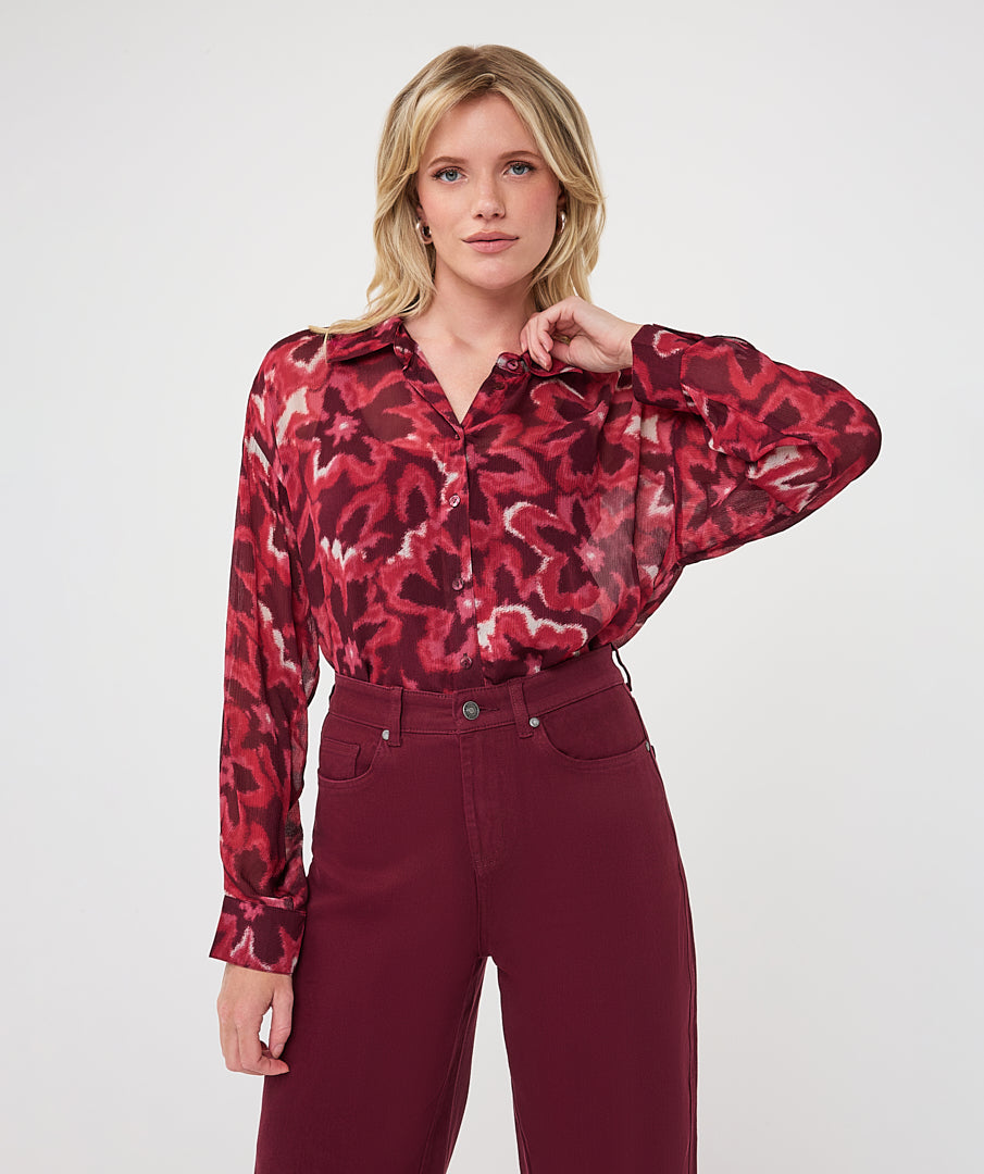 Woman wearing a burgundy and red abstract print button-front blouse with a collared neckline and long sleeves, styled with high-waisted burgundy pants.