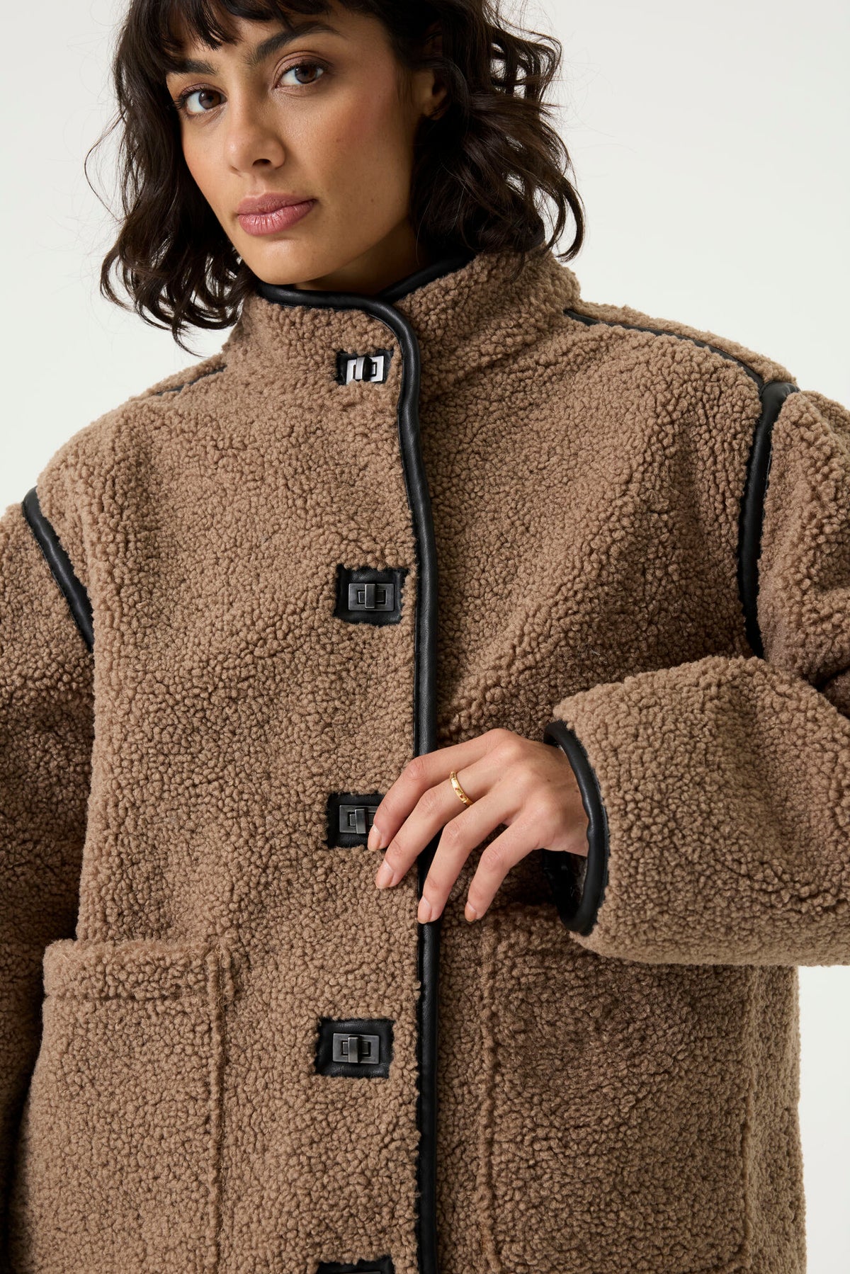 Woman wearing a brown teddy fleece jacket with a high collar, toggle closures, patch pockets, and blue jeans,