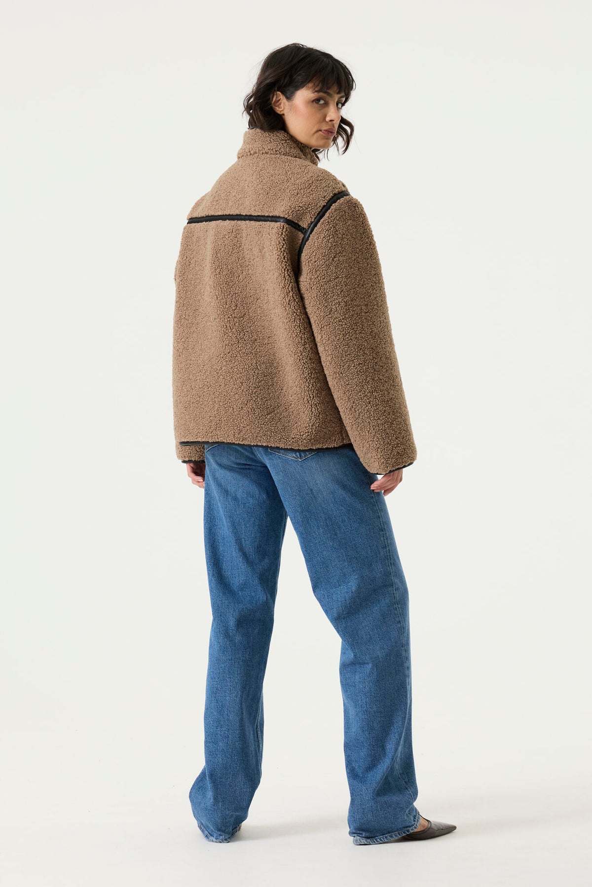 Back view of a woman wearing a brown teddy fleece jacket with a relaxed fit, high collar, contrast trim detailing, and blue jeans.