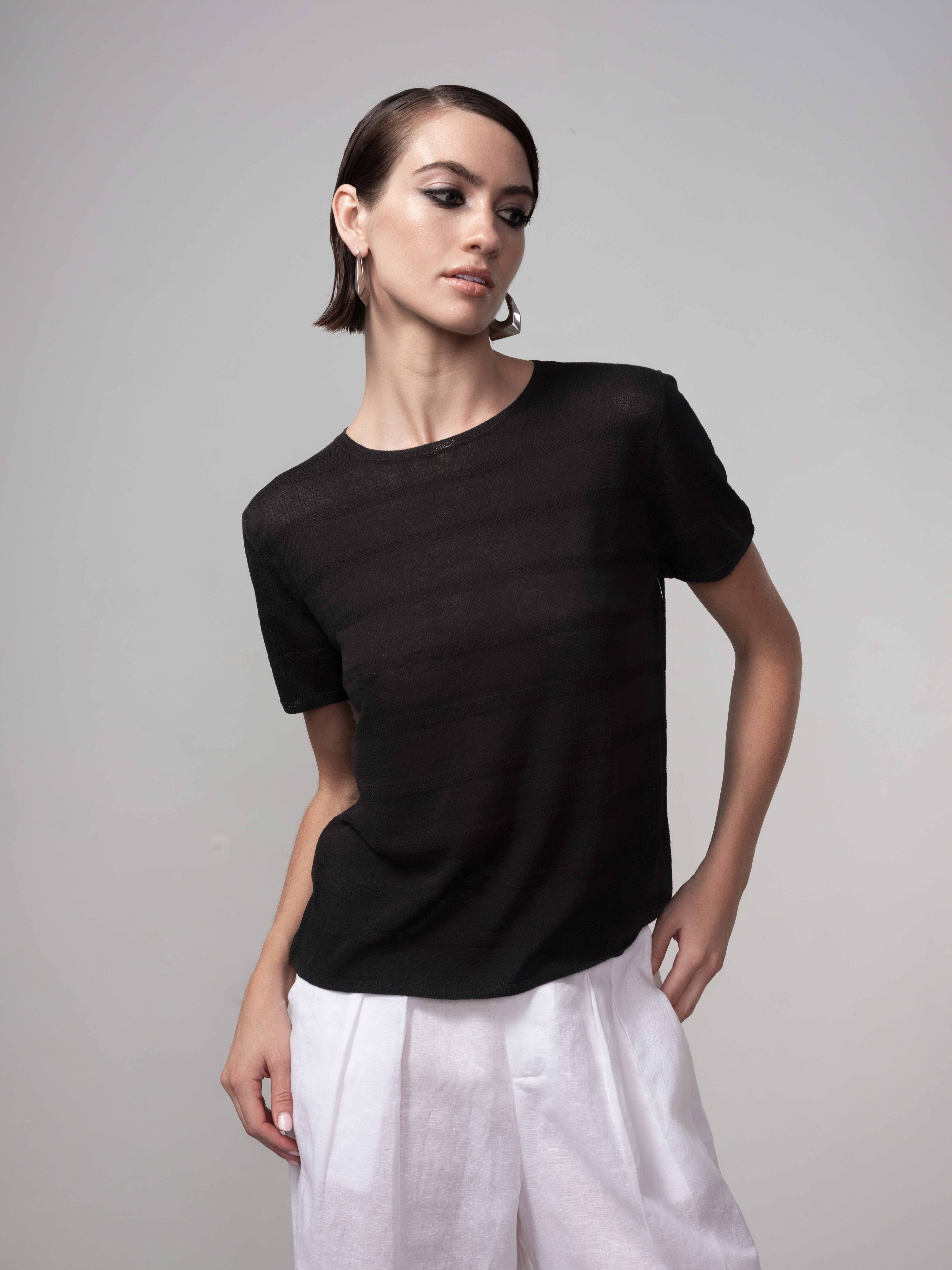 Woman wearing a black lightweight sheer knit short-sleeve sweater with a relaxed fit, paired with white wide-leg pants, shown in a studio setting.