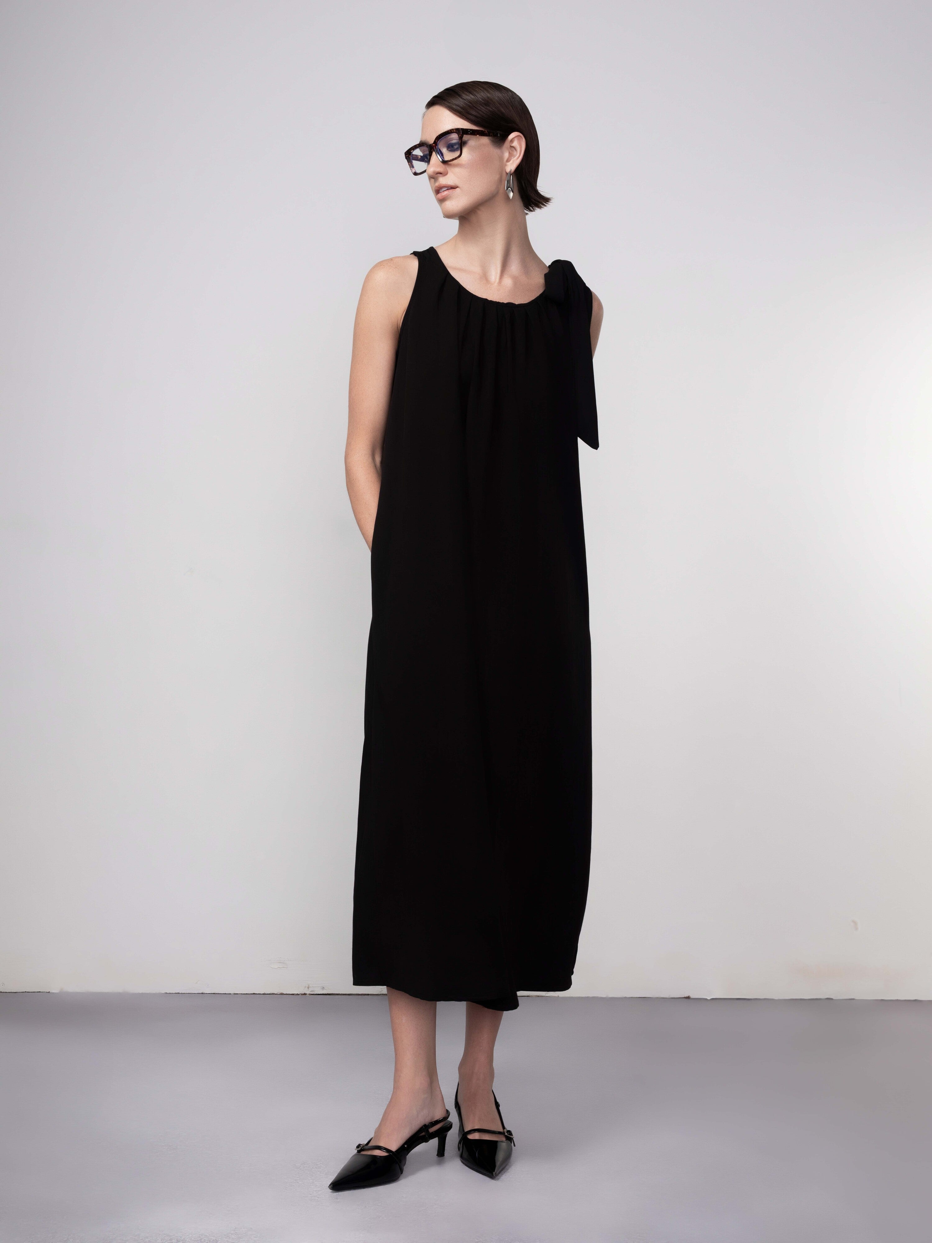 Woman wearing a black sleeveless midi dress with gathered neckline and tie-shoulder detail, paired with pointed-toe flats and glasses.