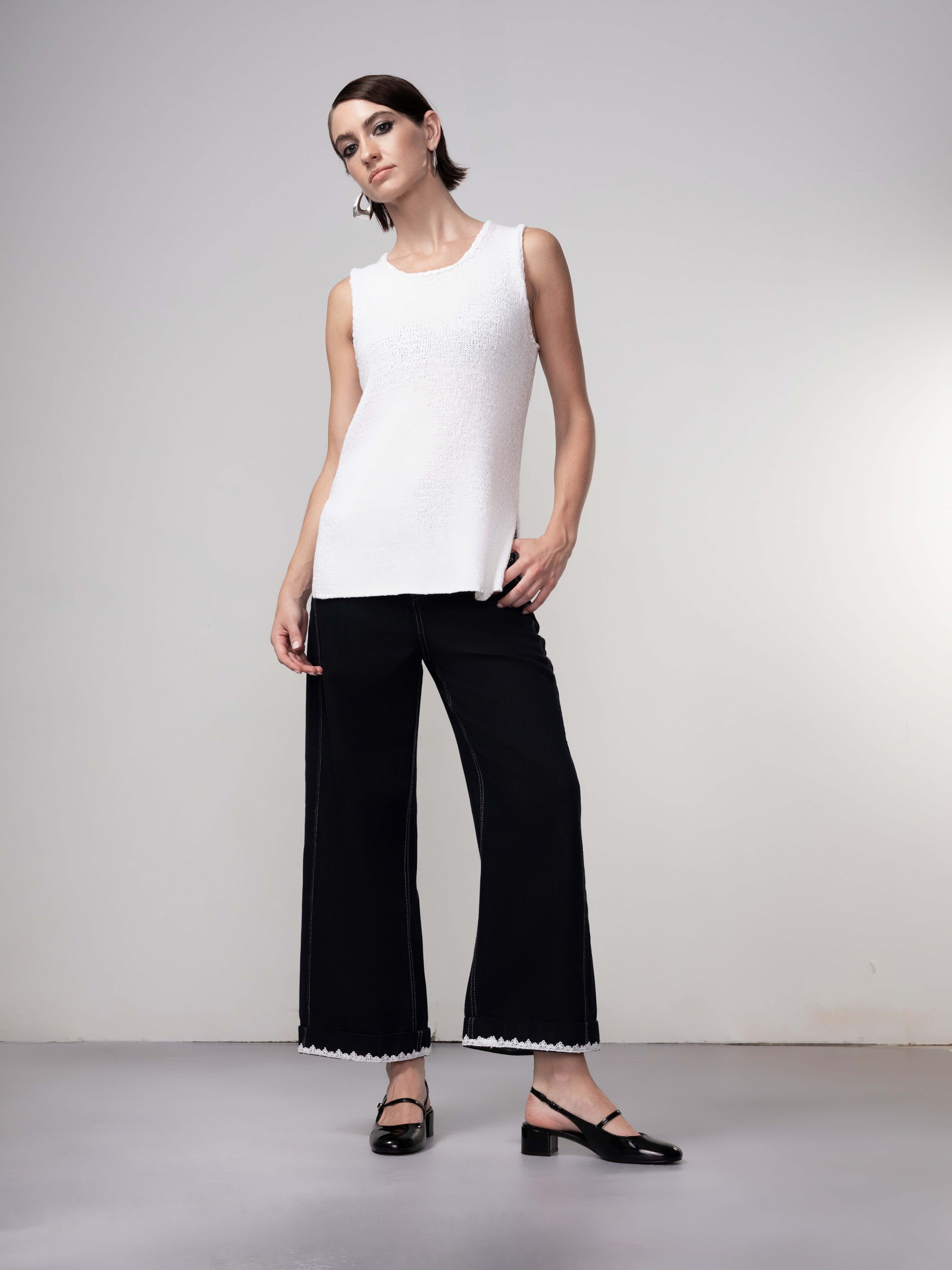 Full-length view of a woman wearing a white sleeveless knit shell top with wide-leg dark jeans and flat shoes, styled in a relaxed, modern outfit.