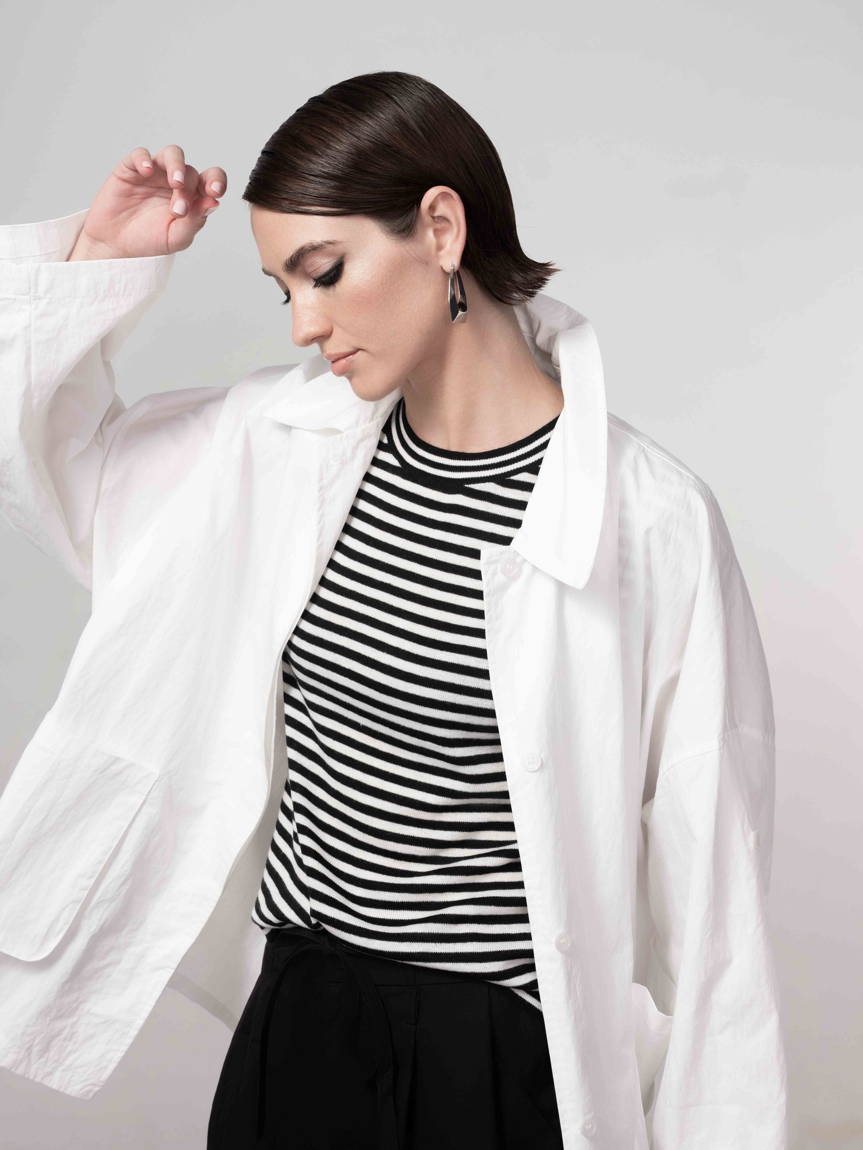 Model wearing a white lightweight jacket layered over a black and white striped top, styled with dark bottoms in a studio setting.