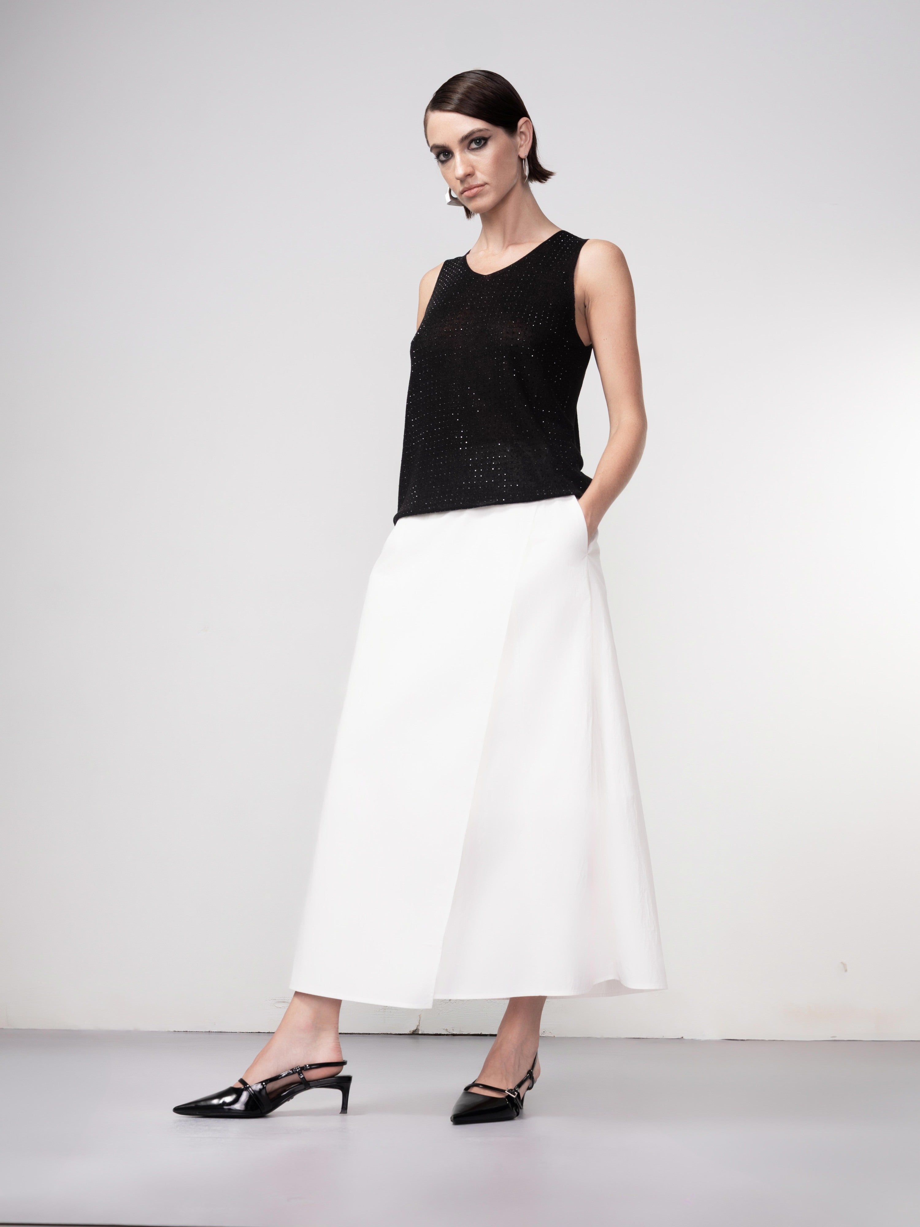 Model wearing a black sleeveless knit tank top styled with a mid-length white skirt and black slingback heels, posing against a neutral studio backdrop.