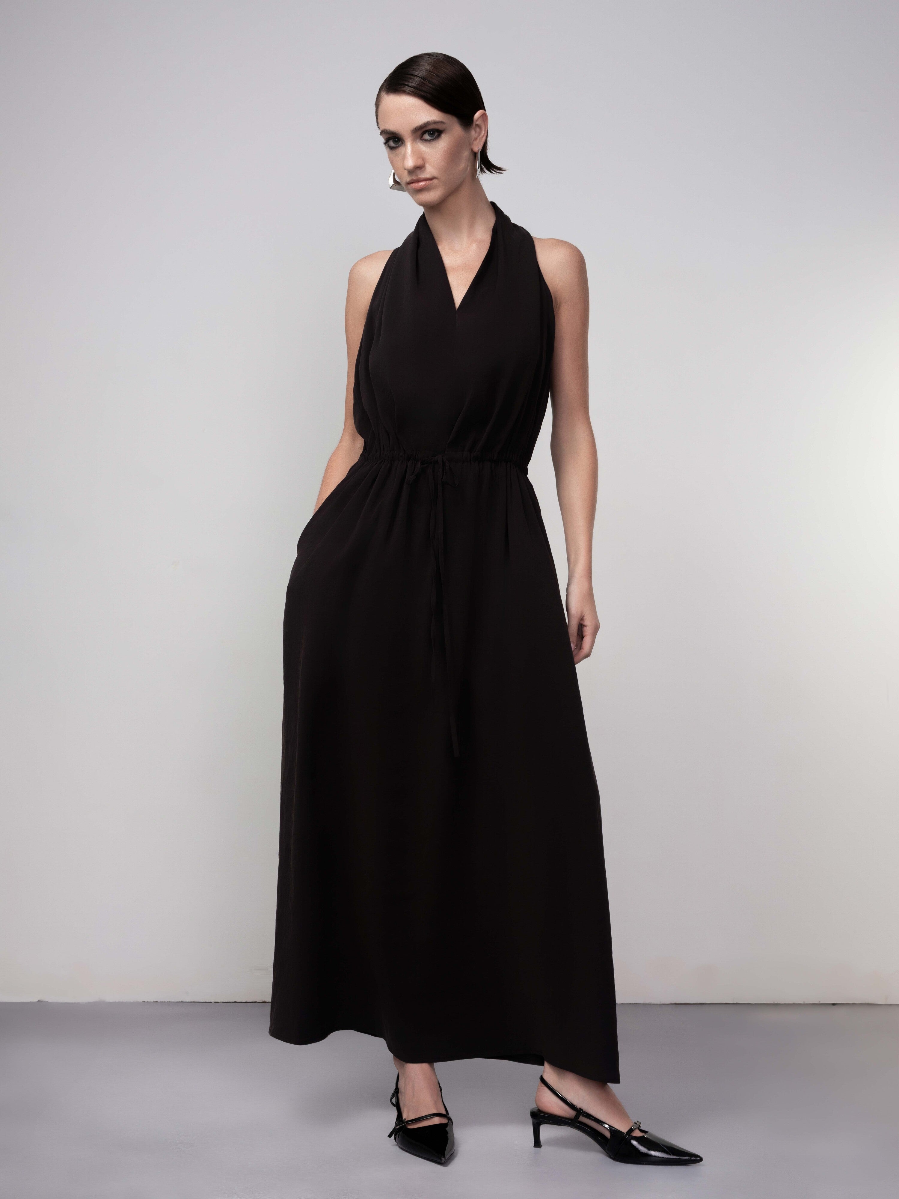 Woman wearing a black halter-neck midi dress with a relaxed draped silhouette, elasticized waist, and fluid skirt, paired with pointed slingback heels against a neutral studio background.