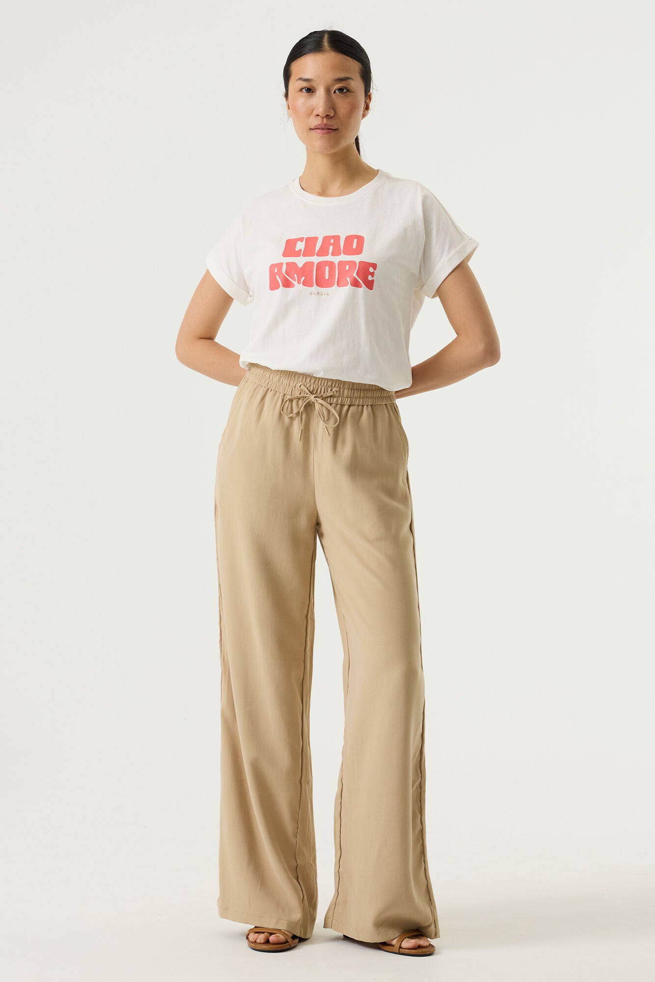 Woman wearing a white relaxed fit graphic tee with “Ciao Amore” text in pink, paired with beige wide-leg drawstring pants and flat sandals