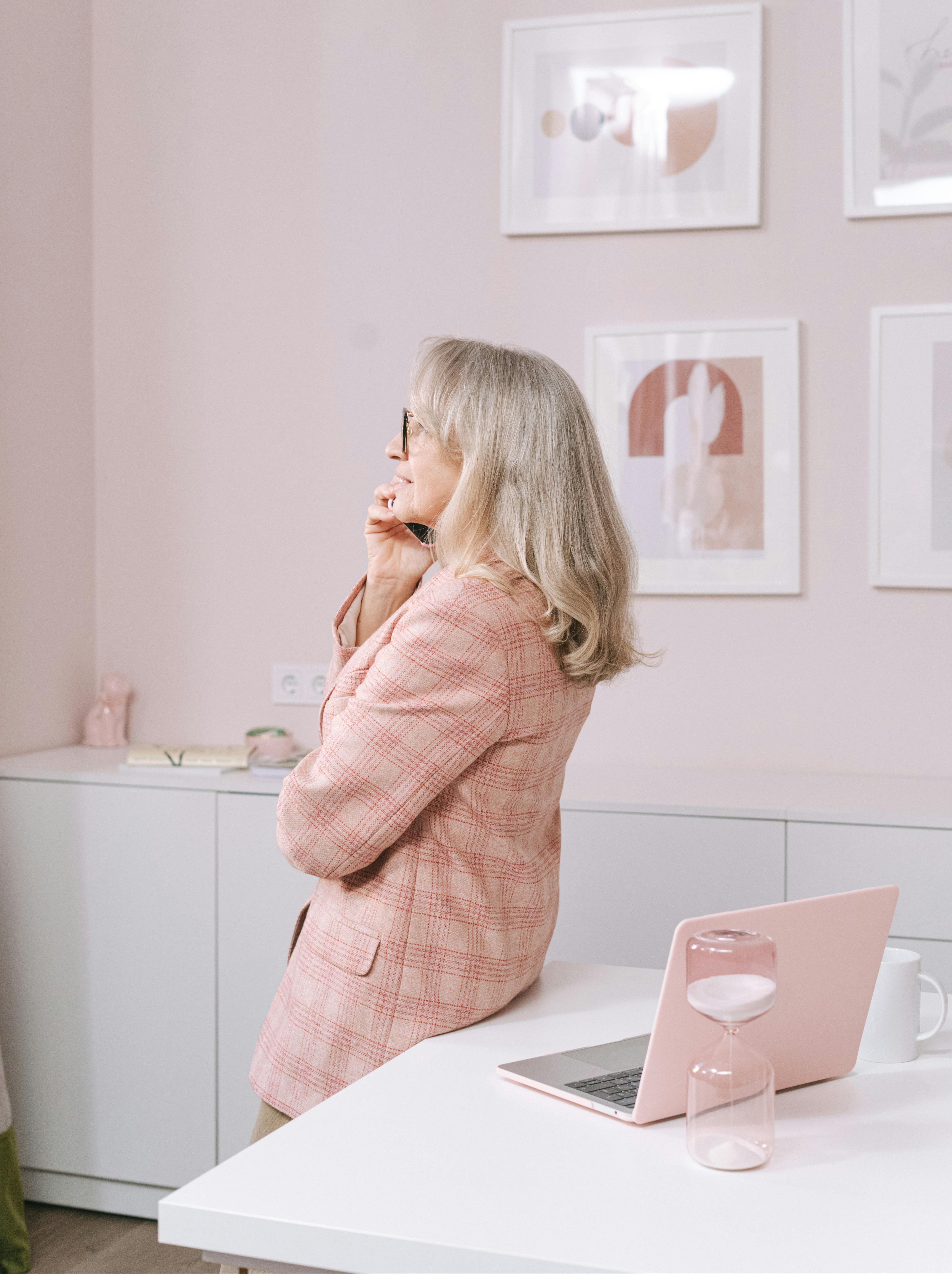 Woman in a pink coat sitting at a desk with a laptop and pink hourglass, against a light pink wall with framed pictures.