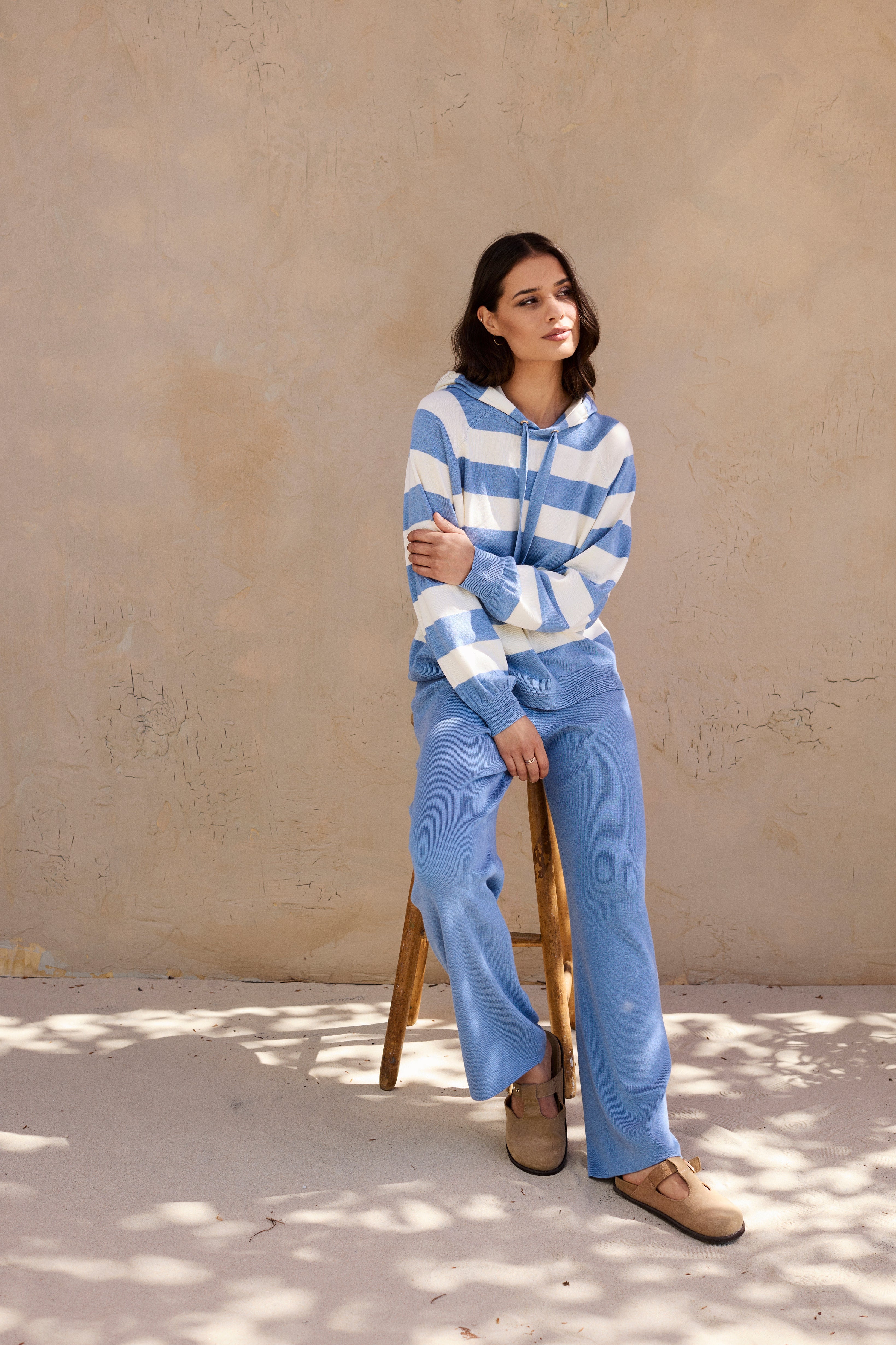Woman wearing a blue and white striped sweater and pants sitting on a wooden stool against a beige wall.