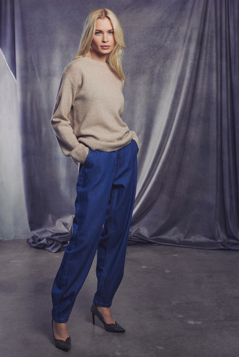 Model in sweater and tapered barrel-leg jeans on a gray backdrop, showcasing a modern casual style.
