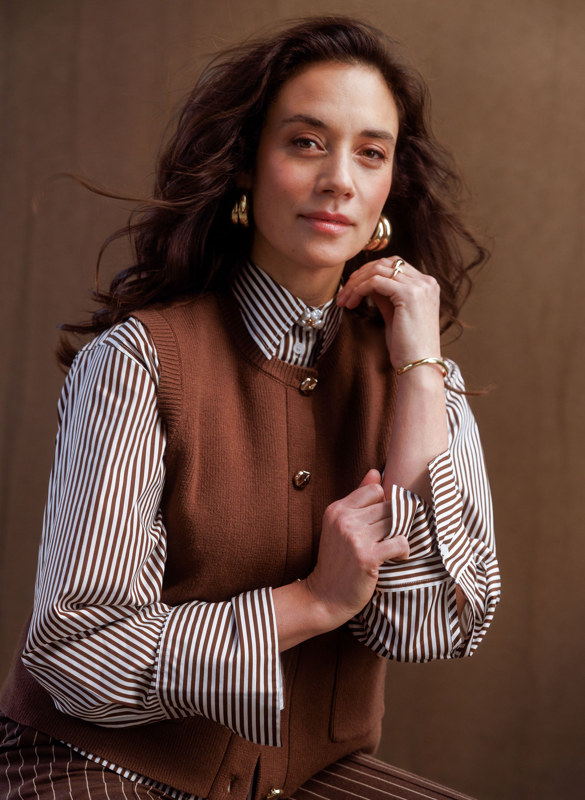 Woman wearing a brown knit button-front vest over a brown-and-white striped long-sleeve blouse, styled with coordinating striped trousers, featuring gold-tone buttons and patch pockets.
