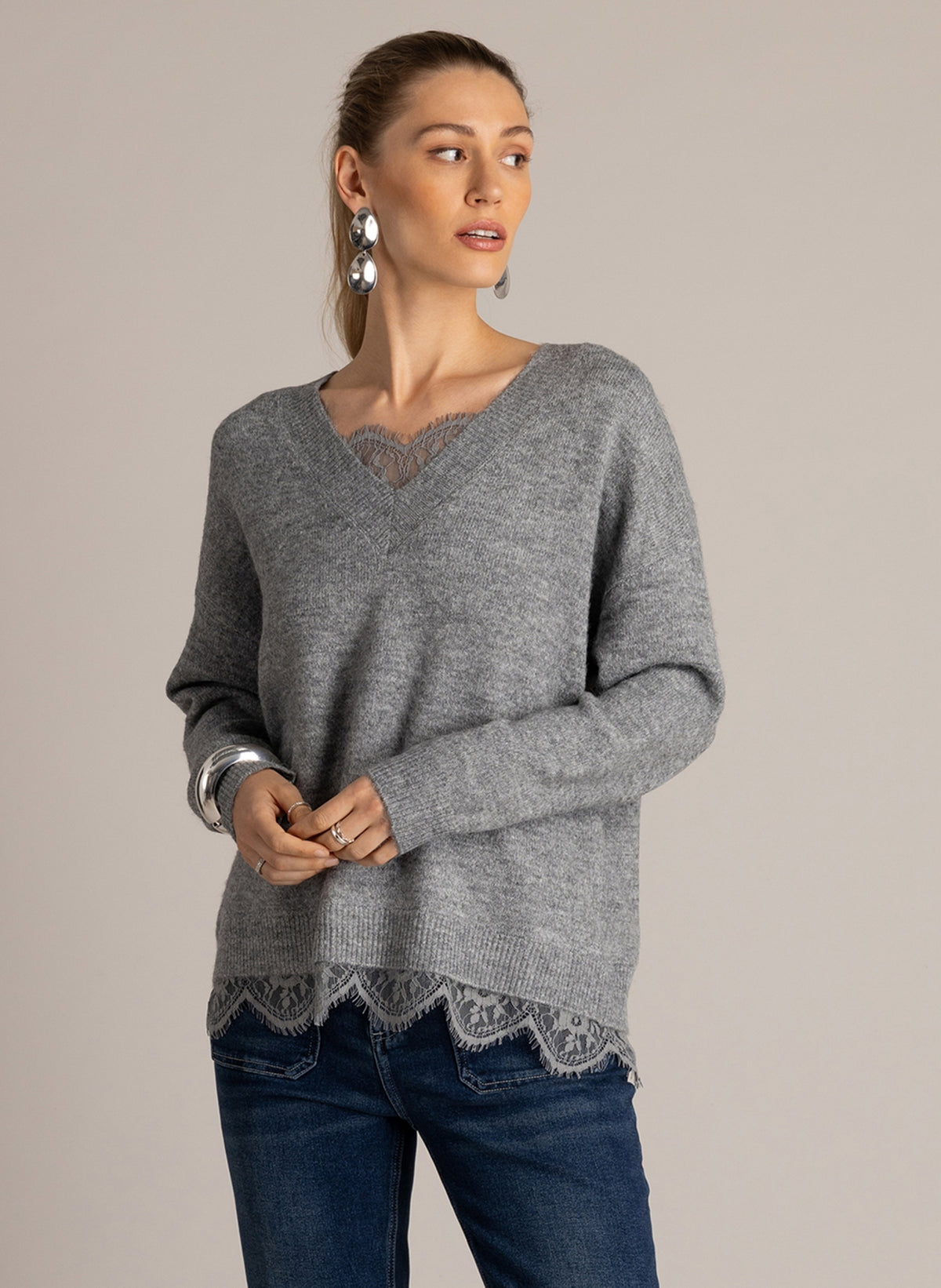 Woman wearing a grey V-neck sweater with lace trim at the neckline, layered over dark wash flared jeans.
