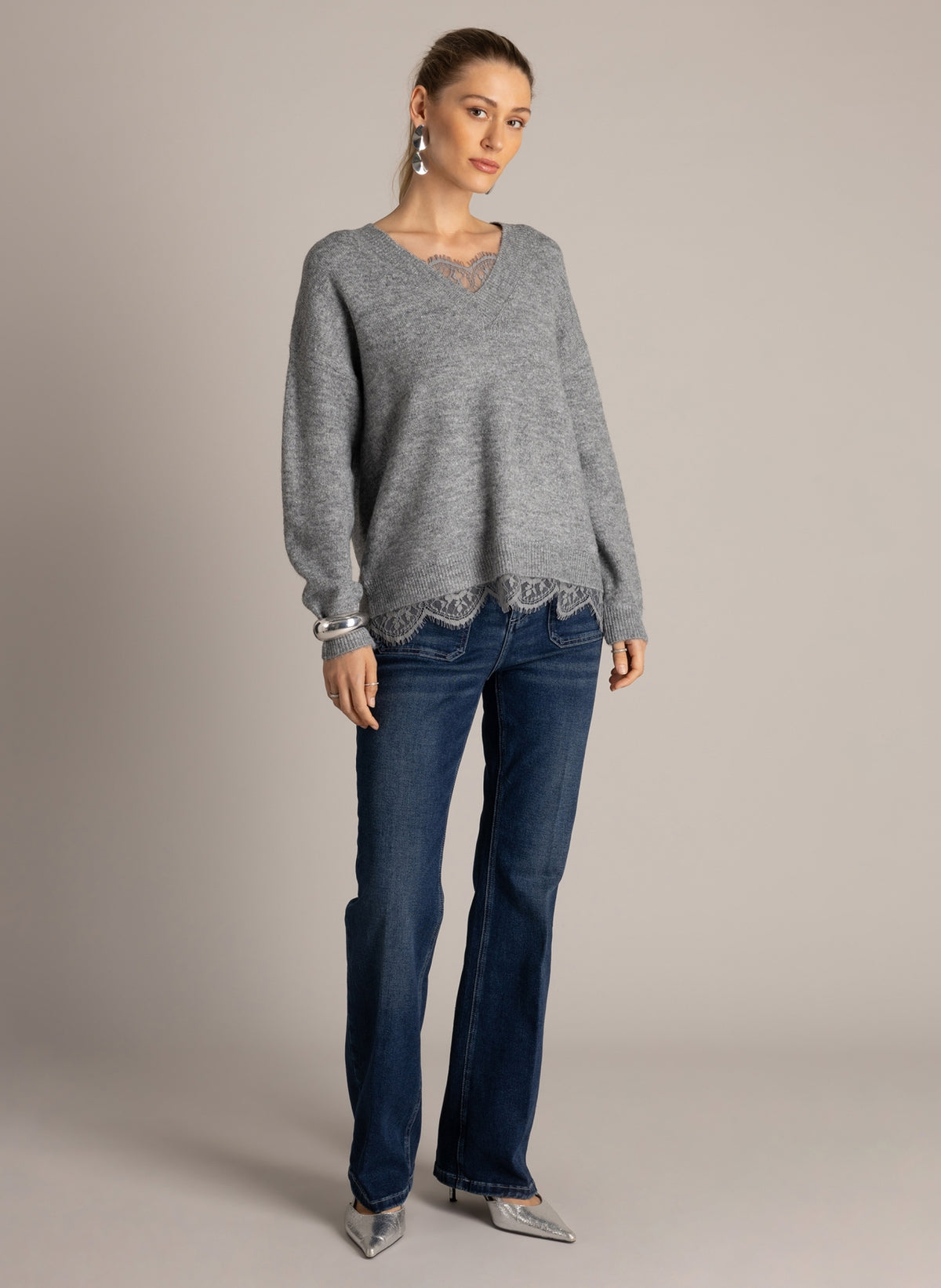 Full-length front view of a grey lace-trim V-neck sweater styled with dark flared jeans and silver heels.
