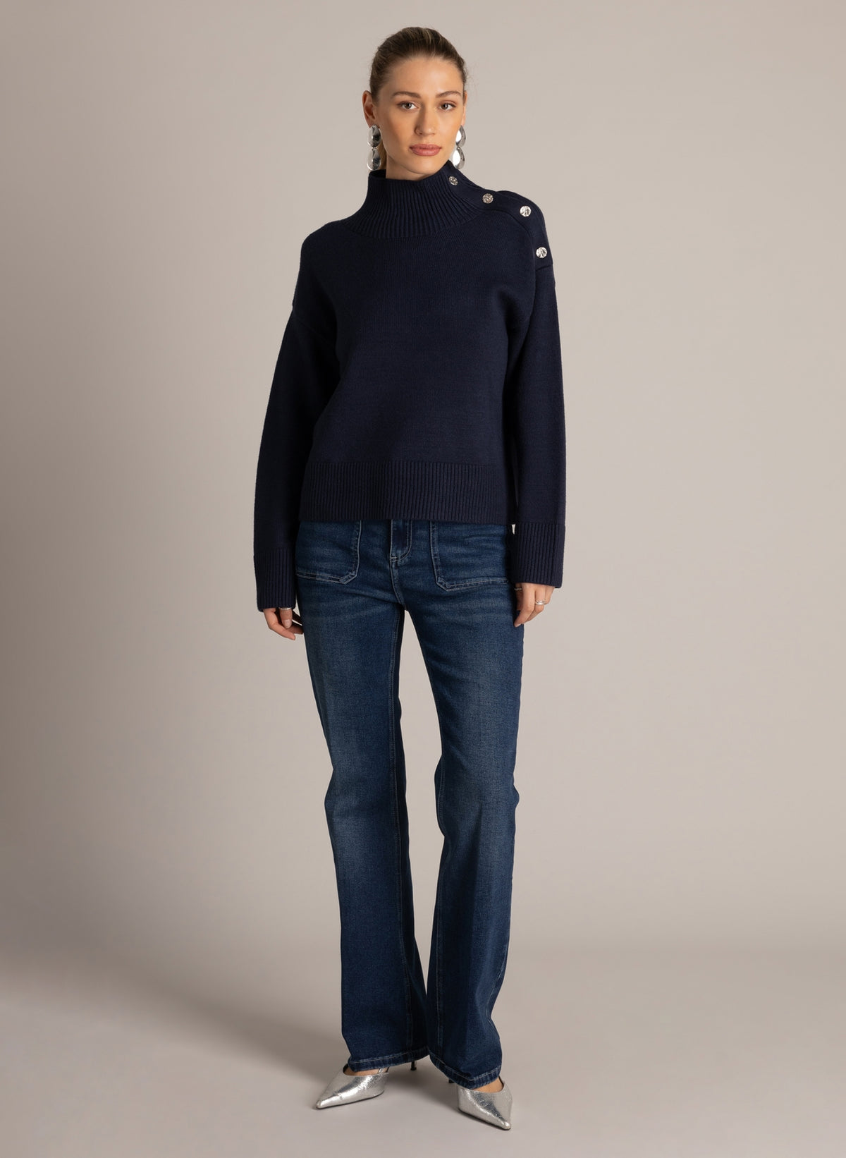 Woman wearing a navy blue button-shoulder sweater with a ribbed mock neckline, styled with dark wash flared jeans.