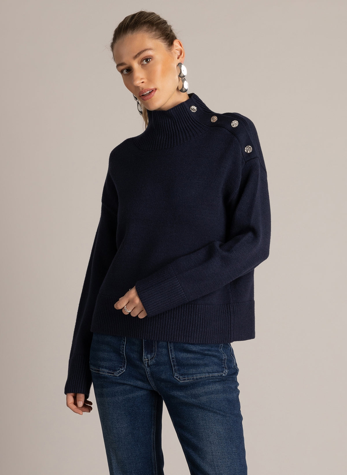 Woman wearing a navy blue button-shoulder sweater with a ribbed mock neckline, styled with dark wash flared jeans.