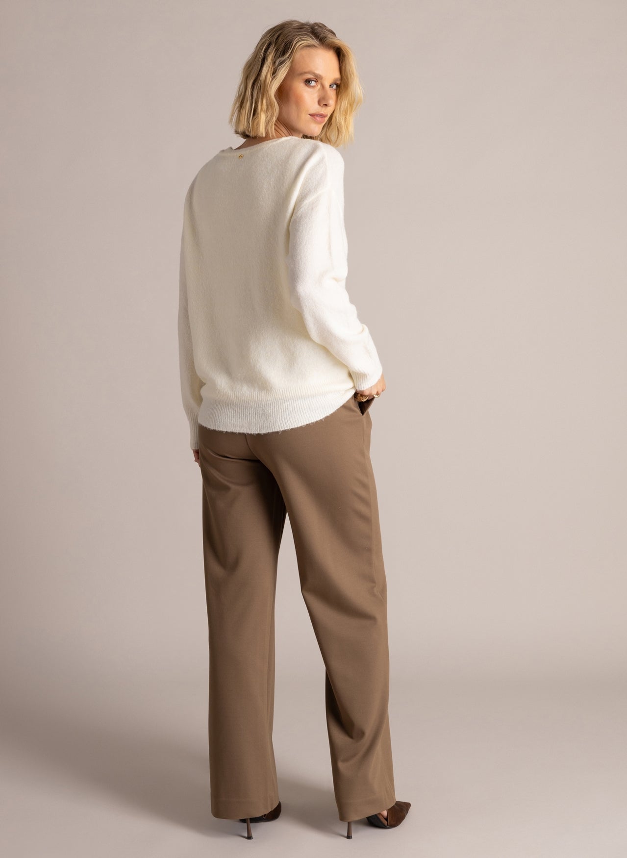 Back view of a woman wearing an ivory long-sleeve V-neck layered sweater with ribbed trim and brown wide-leg trousers.