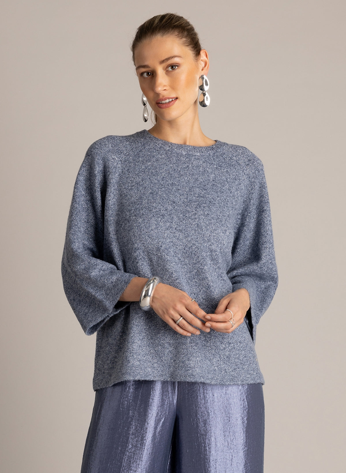 Model wearing a blue-grey textured knit top with three-quarter sleeves and wide-leg, metallic blue-grey trousers.