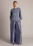  Model wearing a blue-grey textured knit top with three-quarter sleeves and wide-leg, metallic blue-grey trousers.