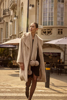 Woman in a long beige coat walking on a city street with buildings and outdoor seating in the background.