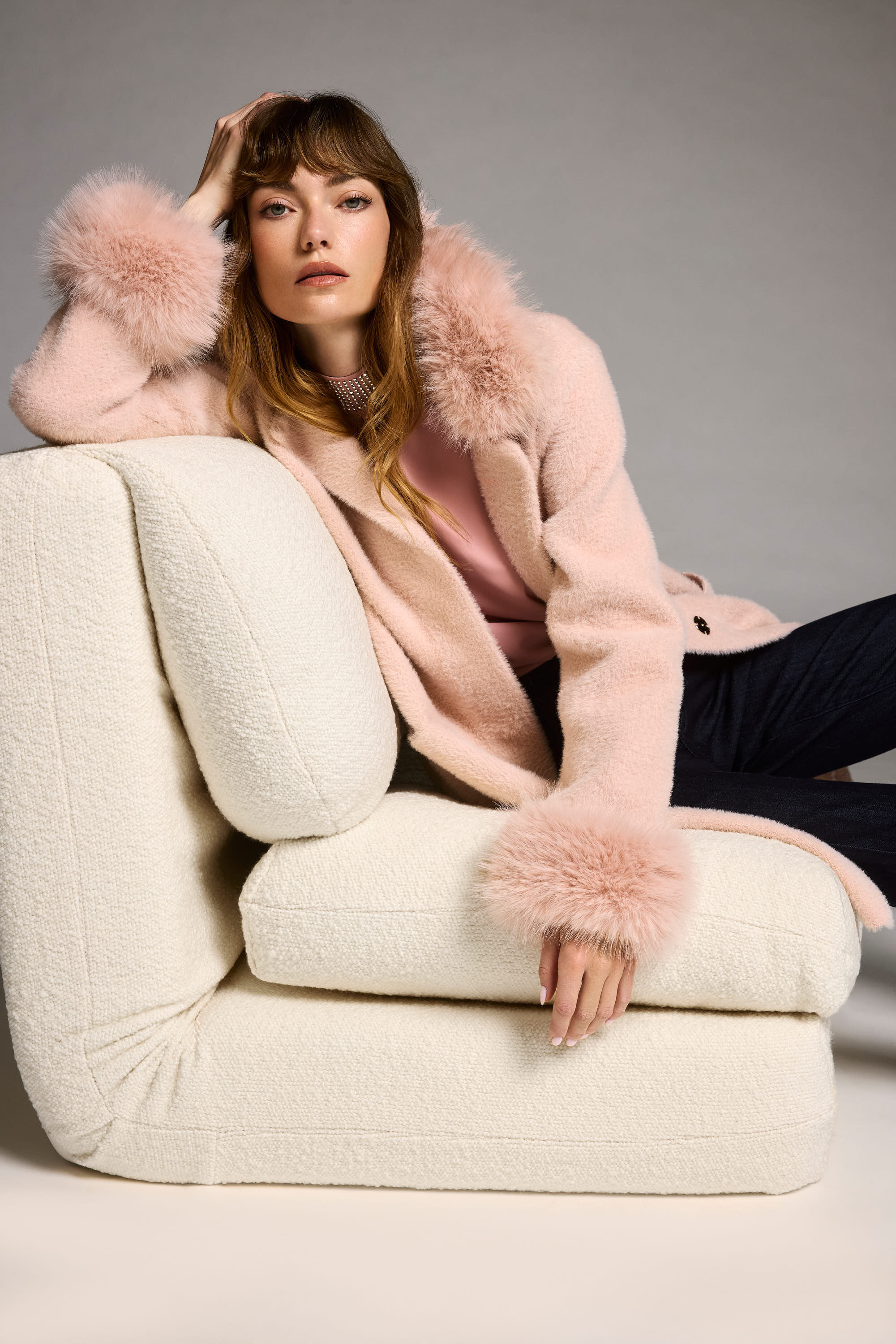 Woman in a pink coat with fur trim sitting on a beige armchair against a gray background