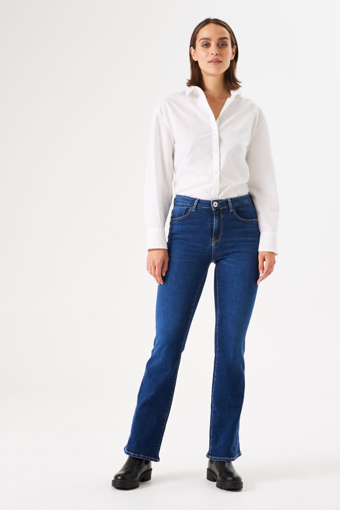 Women's dark blue bootcut jeans with a mid-rise fit, styled with a white button-down shirt and black ankle boots.