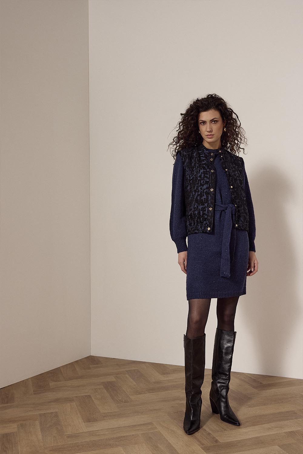 Woman in a navy outfit with a patterned vest standing in a room with beige walls and wooden floor.