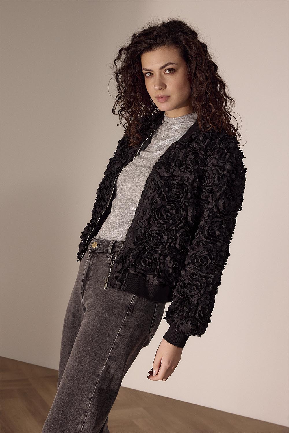Woman wearing a black floral jacket over a gray sweater and jeans against a beige wall.
